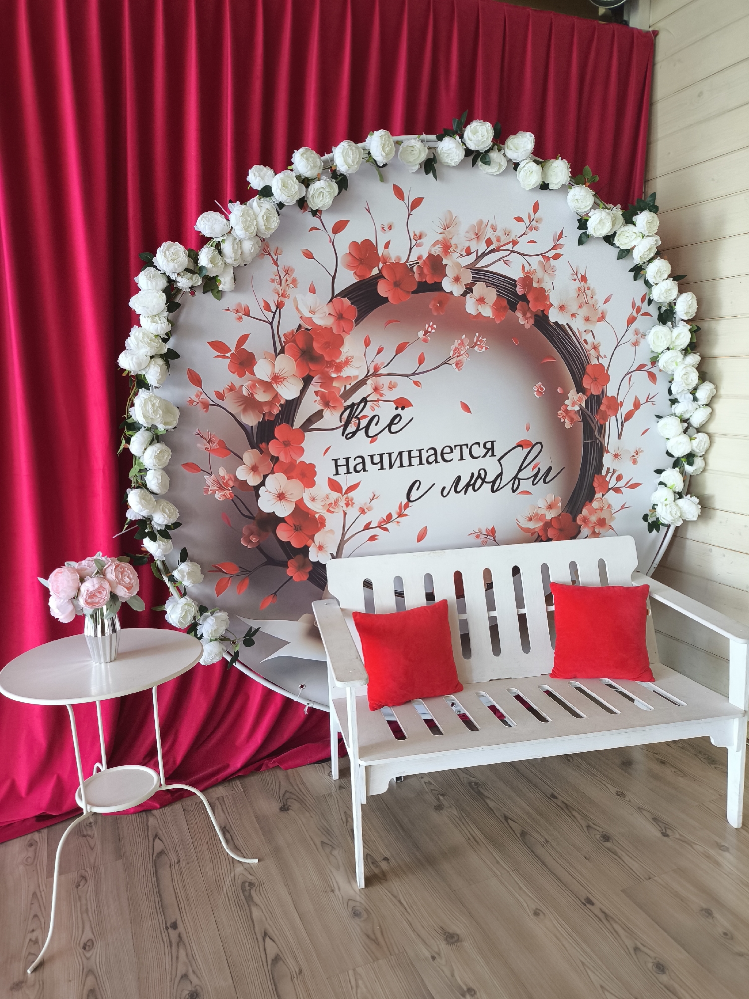 This is a forensic, matter-of-fact description of a scene located in Taldom, Russia. The scene depicts a photo zone with a white wooden bench in the foreground, adorned with two red square cushions. To the left of the bench is a small, white, round side table with a vase containing a bouquet of pink and white flowers. Behind the bench and table, a large, circular backdrop features a floral design with cherry blossoms and the Russian text "Всё начинается с любви" (Everything begins with love). White artificial flowers are arranged in a garland along the edge of the backdrop. To the left of the backdrop, a vibrant fuchsia curtain is draped. The floor is made of light-colored wooden planks. The background wall to the right of the backdrop appears to be made of light-colored wooden planks as well. No people are present in the scene. The lighting appears to be natural or artificial, creating a bright and clear image.