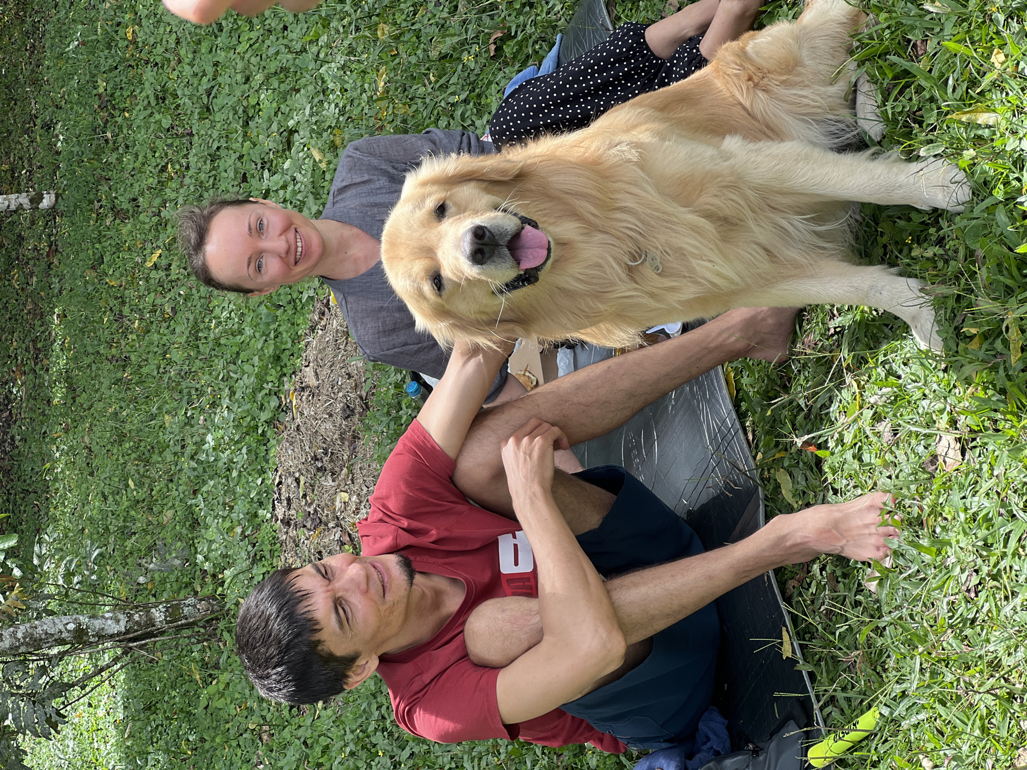 The image shows a woman, a man, and a golden retriever dog in a grassy area, likely in a park or yard. The woman is fair-skinned with short brown hair and is wearing a gray top. She is smiling at the camera. The man is fair-skinned with short dark hair and is wearing a red t-shirt with a white number 9 on it and blue shorts. He is lying on the ground with his legs bent. The dog is lying on top of the man's legs, with its head facing the camera and its tongue sticking out. The dog is wearing a collar. Behind the woman is another person wearing a black and white polka-dotted skirt. The scene is situated in the city of Baturiti, Indonesia. The setting appears to be outdoors with green foliage and good natural lighting. The ground is covered with grass and some loose vegetation. A black mat is visible on the ground beneath the man and the dog.