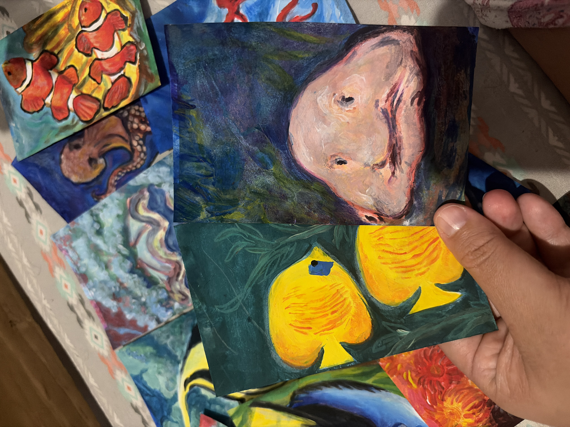 The image shows a collection of small painted canvases or cards, primarily depicting underwater themes. There are paintings of clownfish, an octopus, yellow fish, and abstract ocean scenes. One painting appears to be a portrait of a person with an indistinct expression. A hand holds one of the yellow fish paintings. The setting is a casual, indoor environment, likely a home or studio, as indicated by the patterned bedding in the background. There is no indication of the city of Ciudad Guayana, Venezuela, or any specific location.