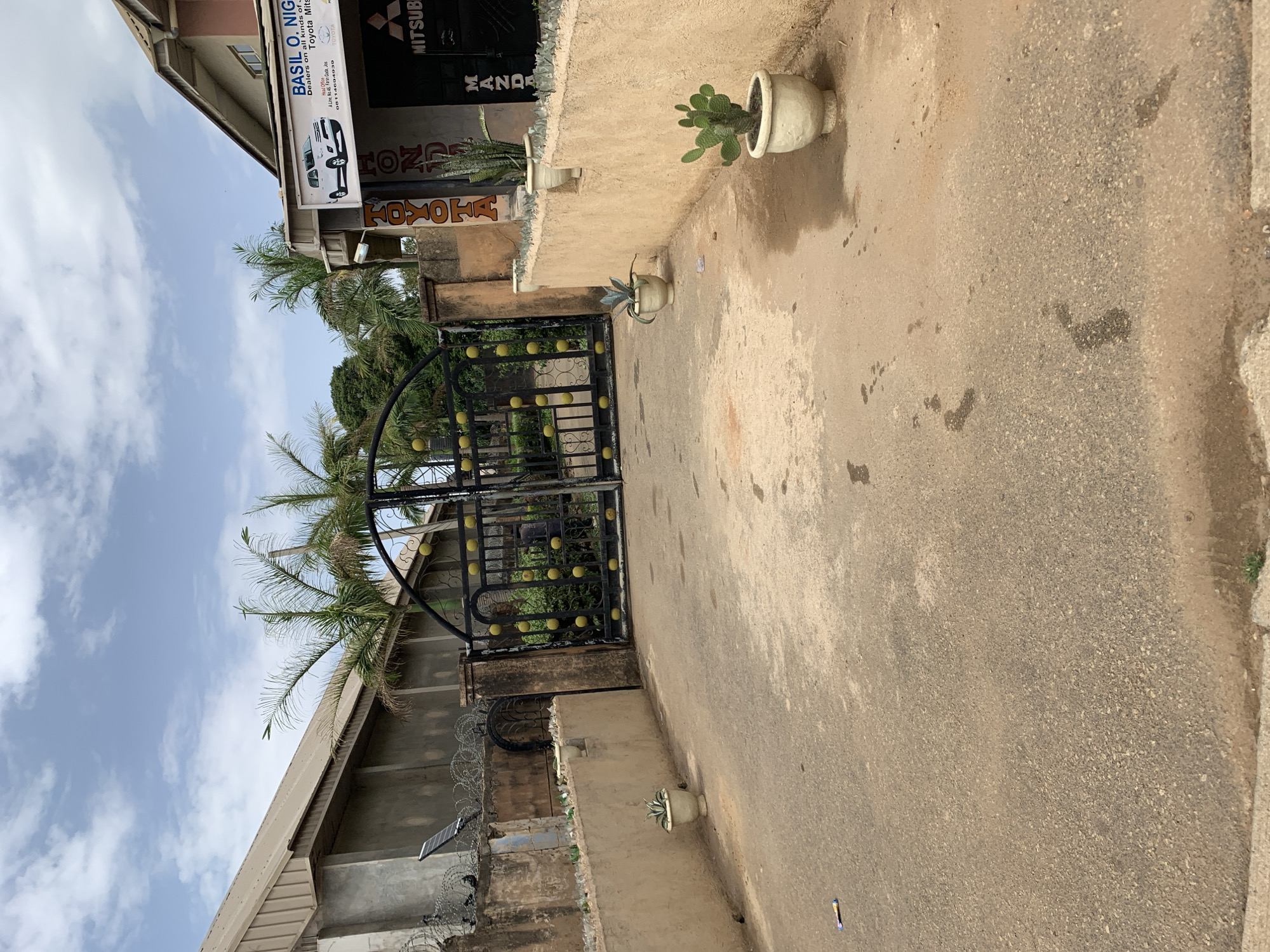 Here's a matter-of-fact description of the image:
The image shows the exterior of a business in what appears to be Rafin Par, Nigeria. The business, "BASIL O. NIG", is a dealer in Toyota, Mitsubishi, Honda, and Mazda vehicles. The building is a two-story structure. In front of the building is a black metal gate with yellow decorative orbs. Several potted plants are visible near the entrance. A concrete driveway or walkway leads up to the building. There is a roof and a wall with razor wire, and a solar panel. The sky is blue with white clouds. The photo has been rotated.
