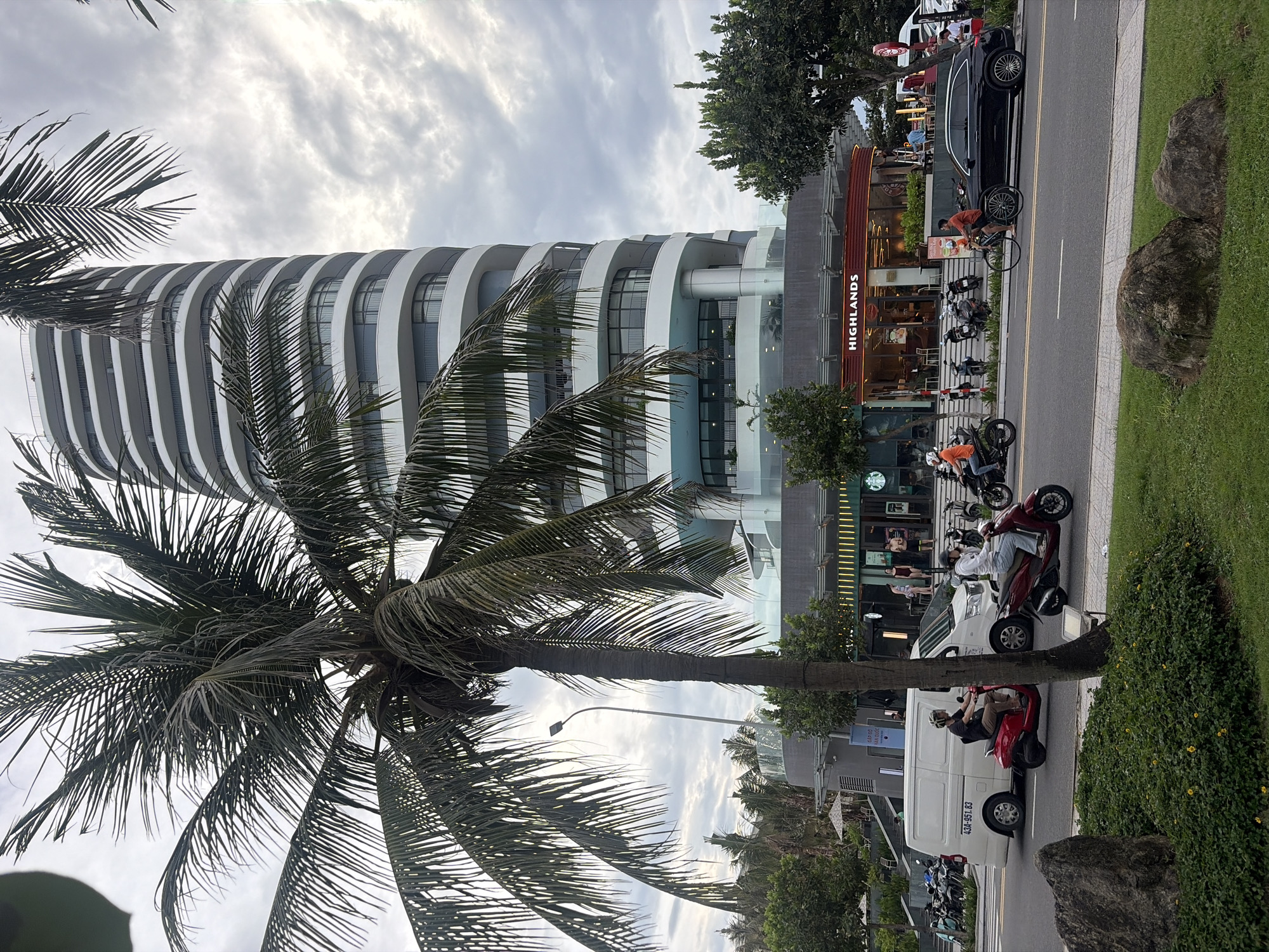 The image captures a street scene in Da Nang, Vietnam, featuring a modern, curved white building with horizontal grey bands and balconies. In the foreground, a large palm tree with lush green fronds partially obstructs the view of the building. A street runs horizontally across the frame, with a white van, several motorcycles, and a black SUV. Two individuals are riding red scooters, and one person is cycling a bicycle. On the ground floor of the building, businesses with signage for "Highlands" and "Starbucks" are visible. The sky is overcast with grey clouds. The overall environment appears to be an urban coastal area, likely a commercial district.