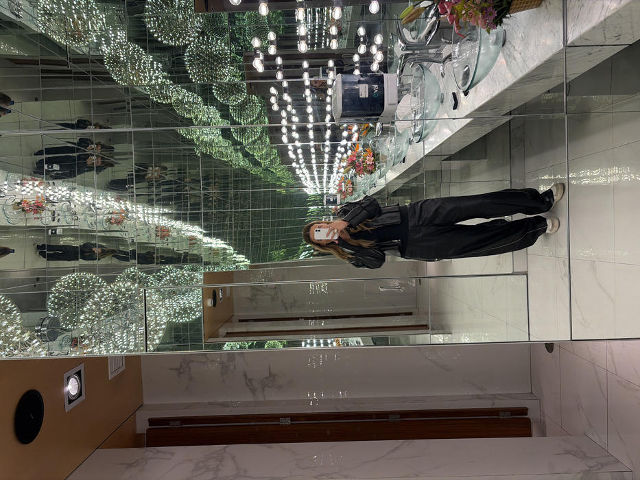 A woman stands in a bathroom, taking a photo with her phone. She is wearing a black jacket and pants. The bathroom has mirrored walls and ceiling, creating an infinity effect. There are sinks, flowers, and lights visible in the reflections. The floor is light-colored tile. A wooden structure and a ceiling light are at the bottom of the image.