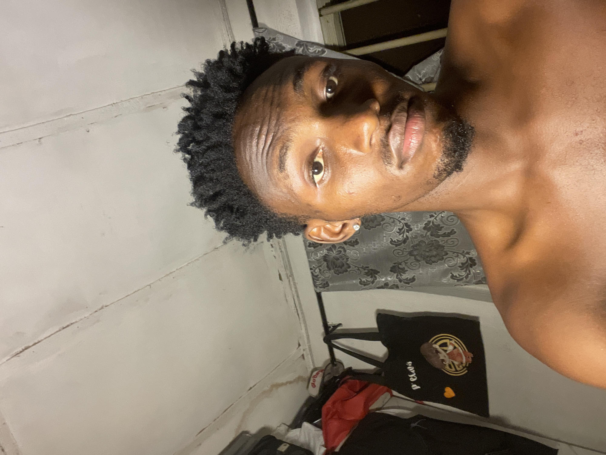 A close-up, low-angle self-portrait of a shirtless Black male with short, curly black hair. He has a goatee and a stud earring in his left ear. The background reveals a sparsely furnished, possibly interior space. The wall is painted white, and appears to have some surface imperfections. A patterned fabric, possibly part of bedding or a curtain, is visible. Also in the background is a black tote bag with gold detailing, as well as other items of clothing. The exact location in Lokoja, Nigeria, is indiscernible from this image.