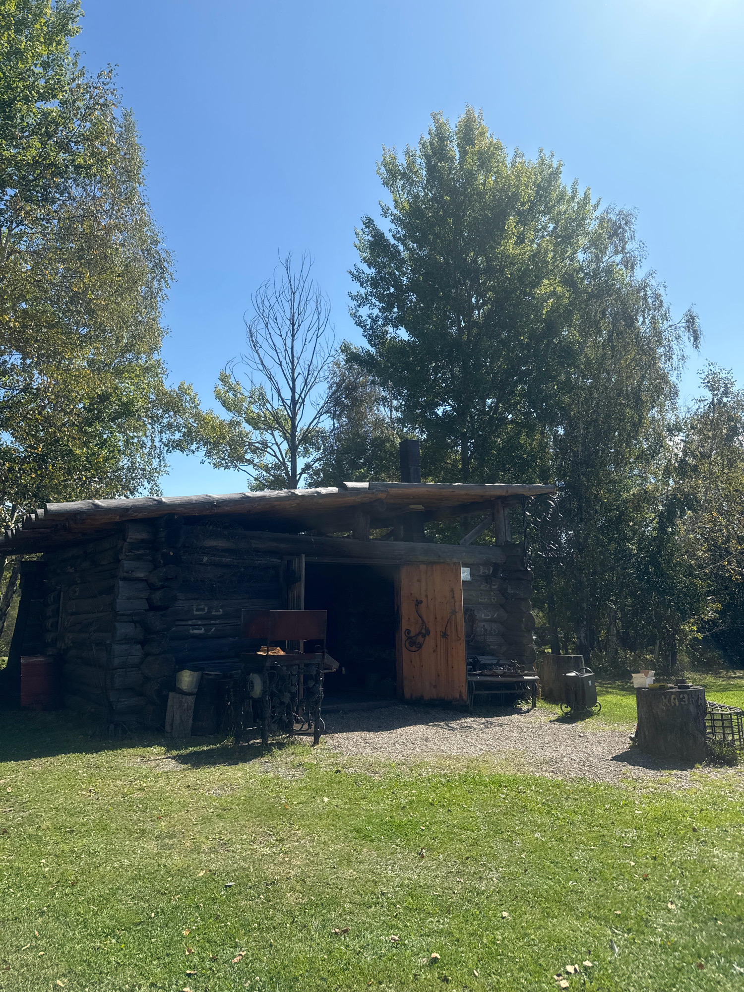 The image depicts a rustic, wooden building situated in the Taltsy Museum, Irkutsk, Russia. The structure is a log cabin with a plank roof. In front of the cabin, there is a wooden door with decorative metalwork. To the left of the entrance, a red barrel and an old metal chair with intricate designs are visible. A metal table with tools and equipment, possibly for blacksmithing, is placed in front of the opening of the cabin. To the right of the cabin, there is a tree stump with some inscription on it and a metal grate. The building is surrounded by a grassy area and trees. The sky is clear and blue. The overall scene suggests an outdoor historical exhibit or a reconstructed traditional dwelling.