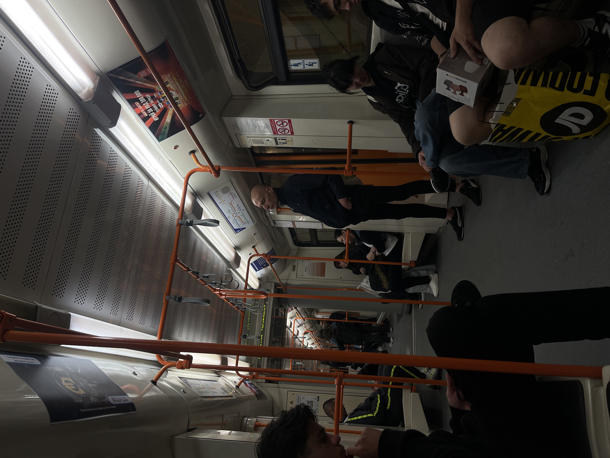 The image depicts the interior of a public transportation vehicle, likely a subway car or train, in Sofia, Bulgaria. The seating is occupied by passengers, some sitting and others standing. One individual is holding a box labeled "ACDECOR," while another has a yellow shopping bag labeled "FOREVER FASHION." An advertisement for PASSET BANK, advertising promotional conditions until September 30, is visible on the side of the vehicle. Another advertisement promotes "GAMING AWARDS" for over 100,000 people. The vehicle interior has orange handrails.