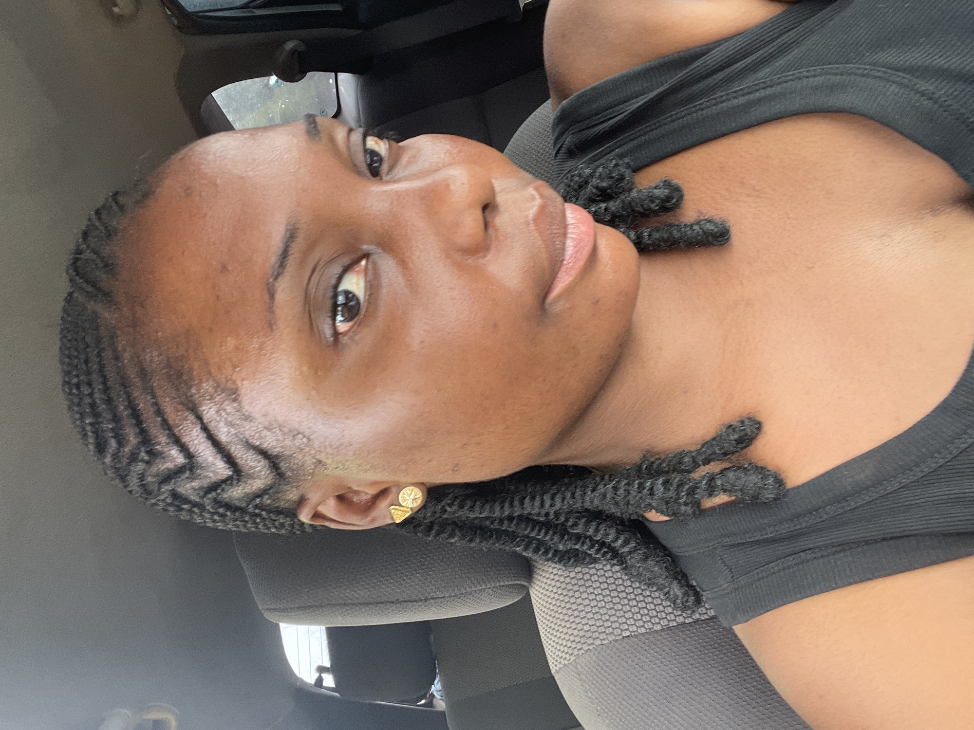 A medium-shot, interior view of a woman in a car. The woman has dark skin and is wearing a black top. Her hair is styled into intricate braids that follow a zigzag pattern along her hairline, with the ends left in textured curls. She has an earring in her left ear. The car seat is grey. The scene takes place in Jos, Nigeria.