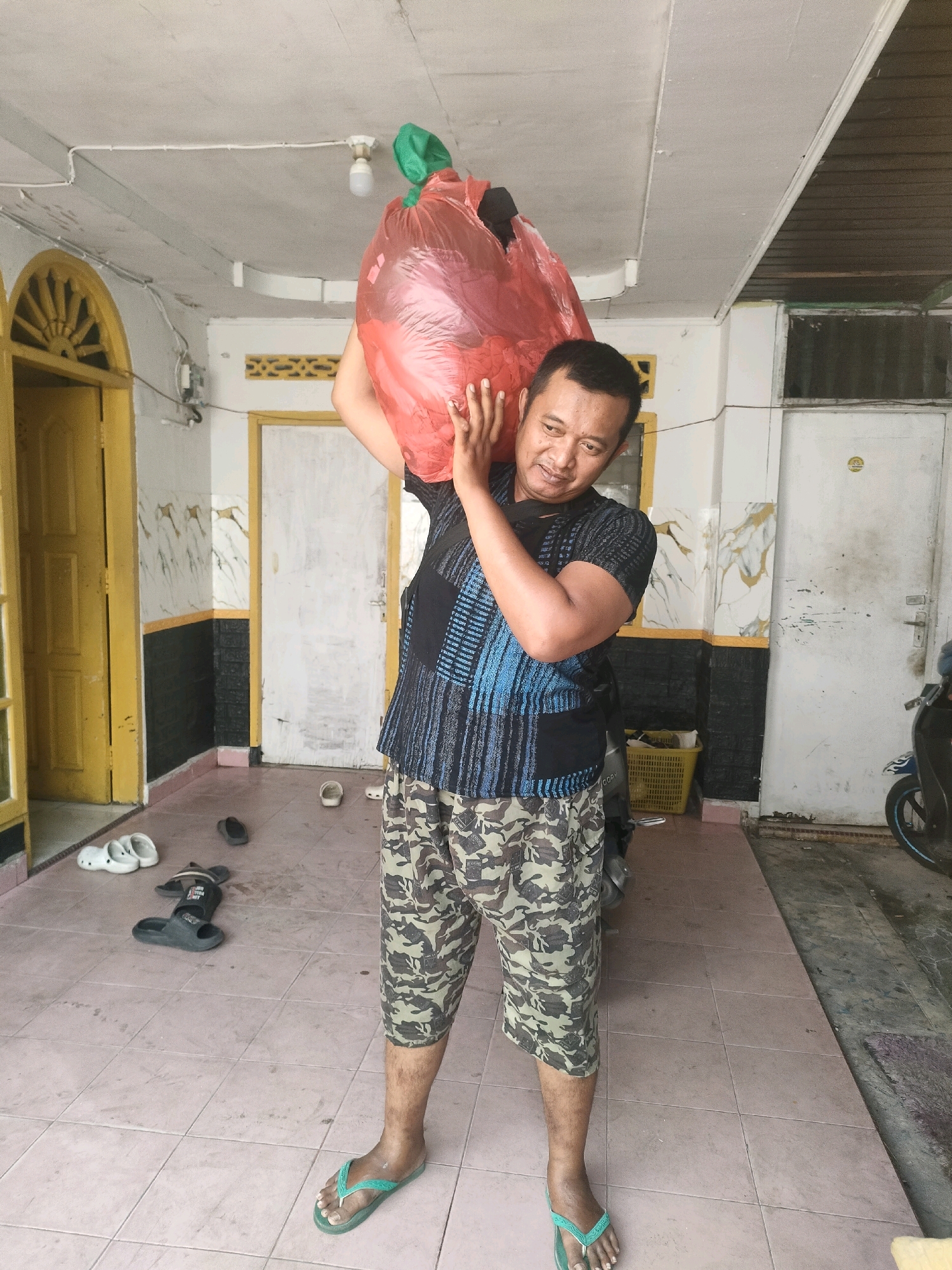 A man is carrying a large red plastic bag filled with what appears to be laundry over his left shoulder. He is standing in what seems to be a covered outdoor area, possibly a porch or a covered walkway, with a tiled floor. To his left, there is a yellow door with decorative elements above it. Further back, there are white doors and a yellow basket. Various pairs of flip-flops and sandals are scattered on the floor. A motorcycle is partially visible on the right side of the image. The walls have a decorative pattern of white and black with gold accents. The man is wearing a patterned t-shirt and camouflage shorts, and green flip-flops. The overall setting suggests a residential area in Pekanbaru, Indonesia, possibly a home or a communal living space.