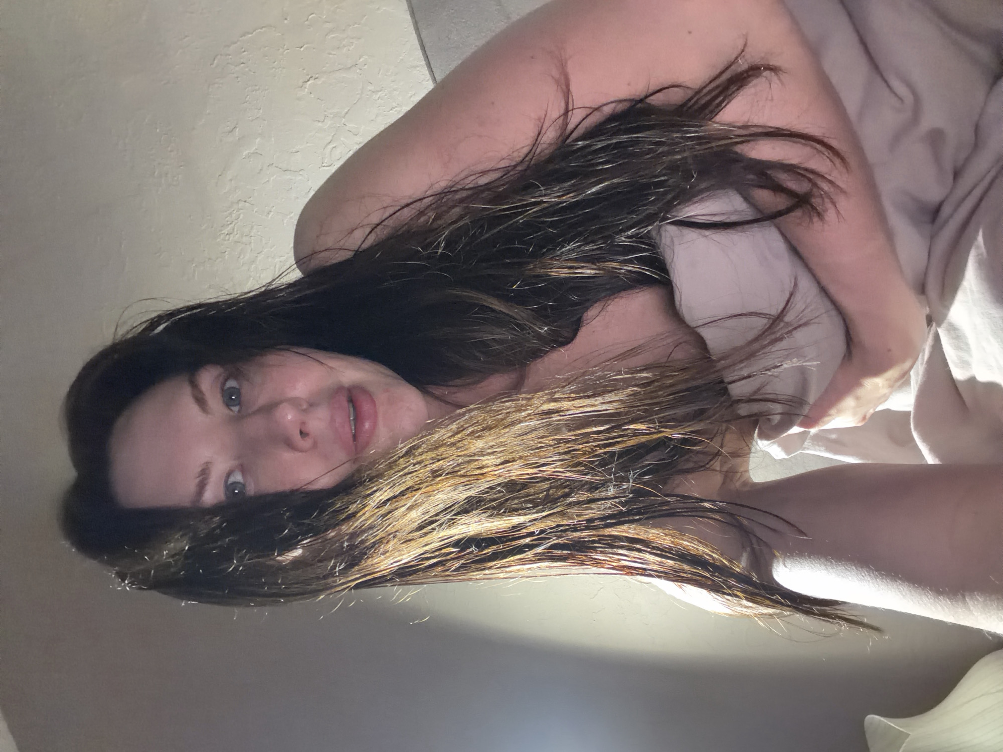 A woman is reclining in bed. She is lying on her back, facing the camera at an angle. Her hair is dark brown with light brown highlights, and it appears to be wet. She is holding a light-colored blanket or sheet close to her body. The background is a textured white wall. The lighting in the room is dim and casts shadows on the wall and her face. The location information is irrelevant, as it does not appear in the image.