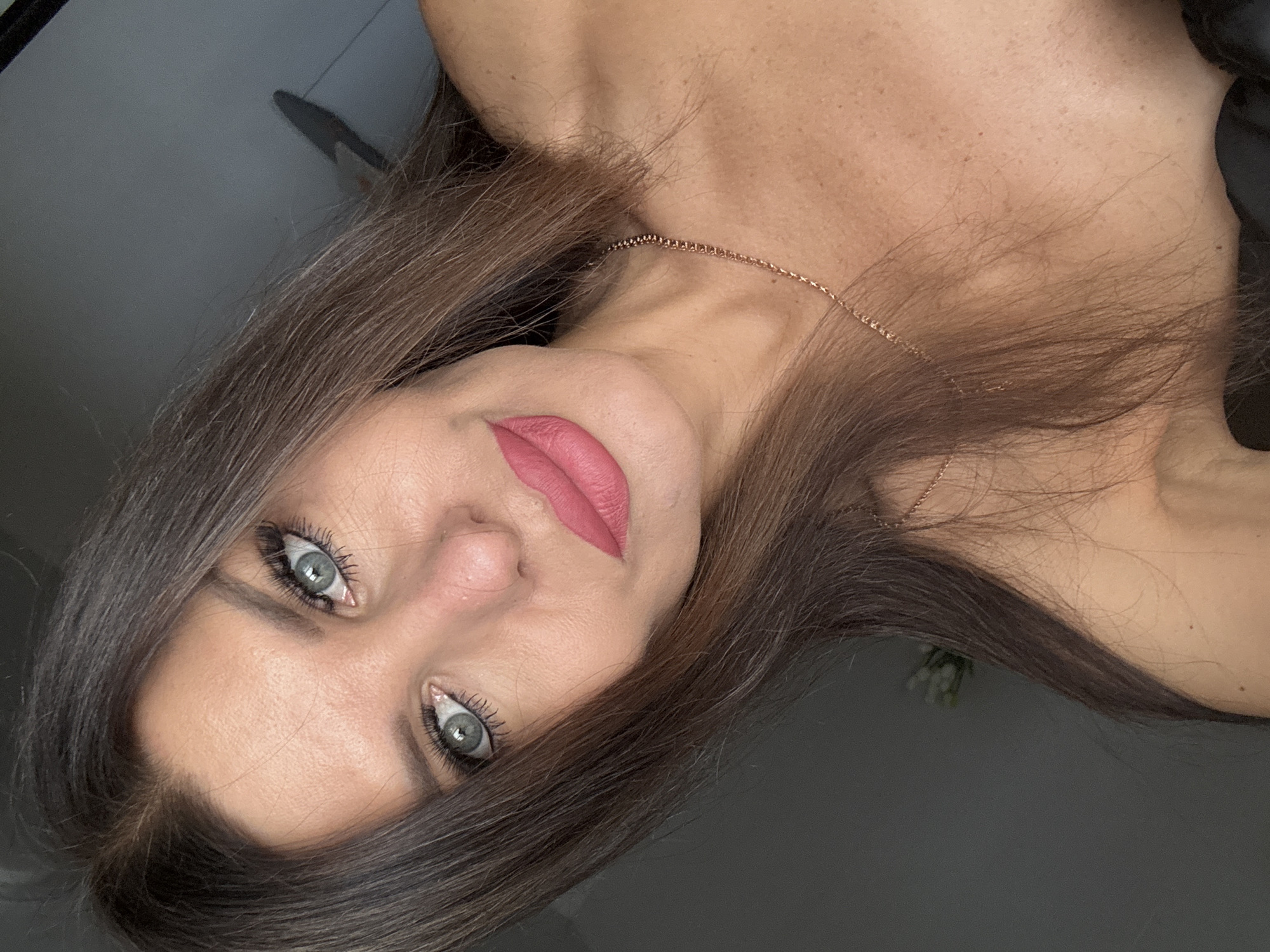 A woman is looking at the camera in a close-up shot. She has long brown hair, light green eyes, and is wearing pink lipstick. She is also wearing a delicate gold necklace. The background is a plain gray wall. There is no indication of the city of Chaikovsky, Russia in the image.