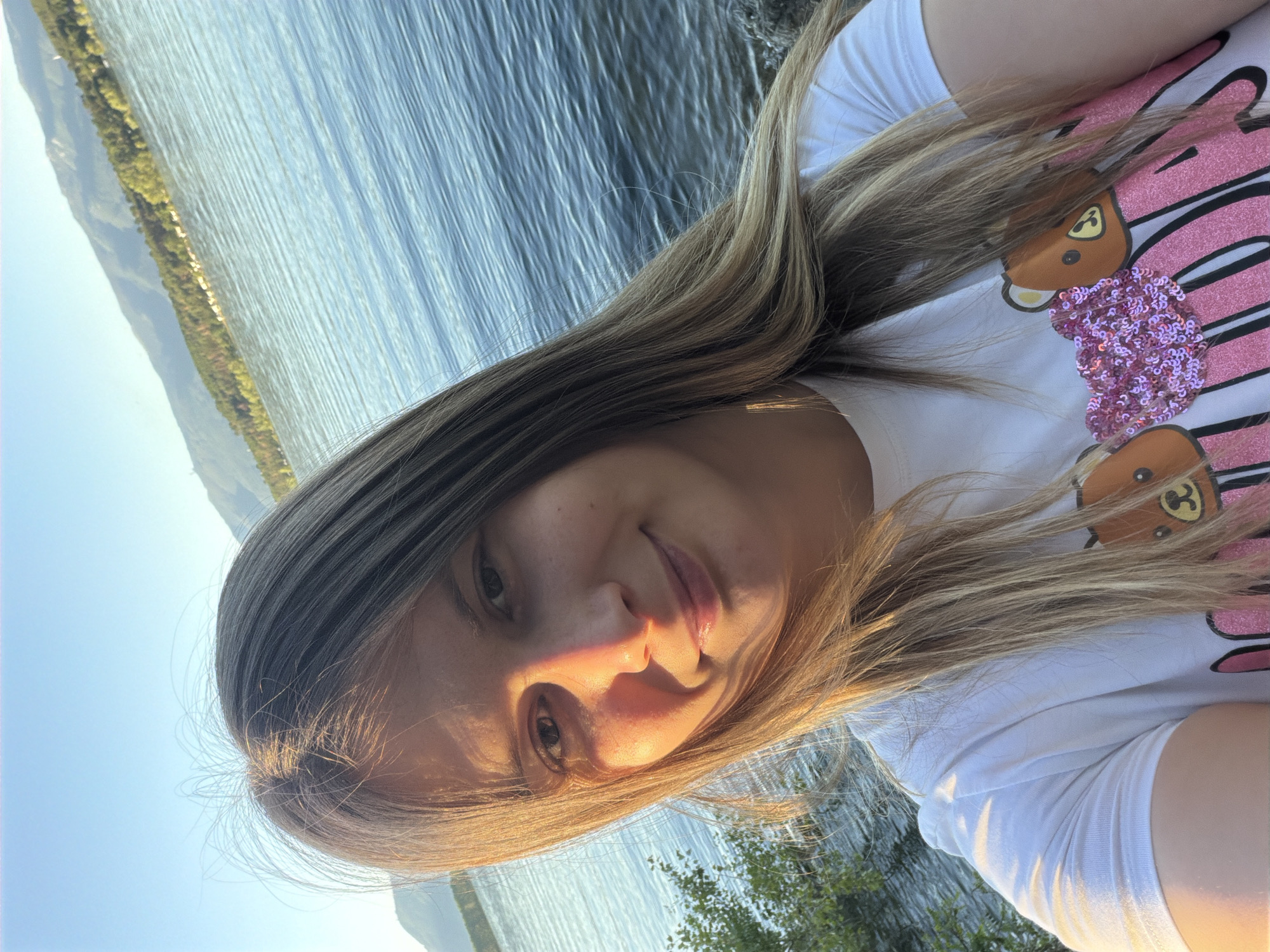 A young woman with long, blonde hair takes a selfie by a lake in Koprinka, Bulgaria. She is wearing a white t-shirt with a graphic of two cartoon bears and pink sequins. The sun is setting, casting a warm glow on her face and the water. Mountains covered in trees are visible in the background.