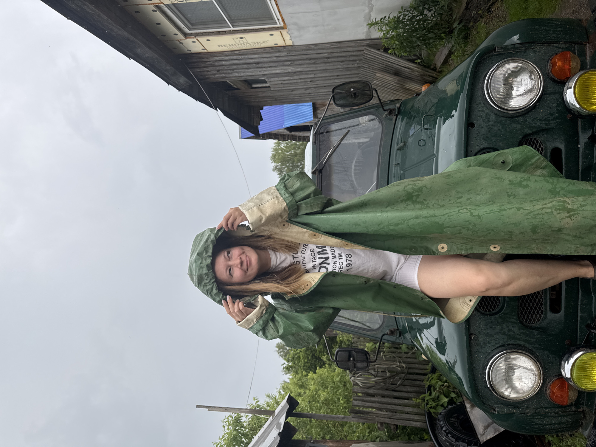 A blonde woman in a green raincoat and white shorts stands on the hood of a green vintage military vehicle. The vehicle has a spare tire mounted on the left side of the hood. The woman is looking to the right of the frame, smiling. Behind her, a wooden building with a partially constructed wall is visible, along with a wooden fence and trees. The sky is overcast, and it appears to be raining lightly. The scene is located in an outdoor, rural setting, possibly a village or countryside.