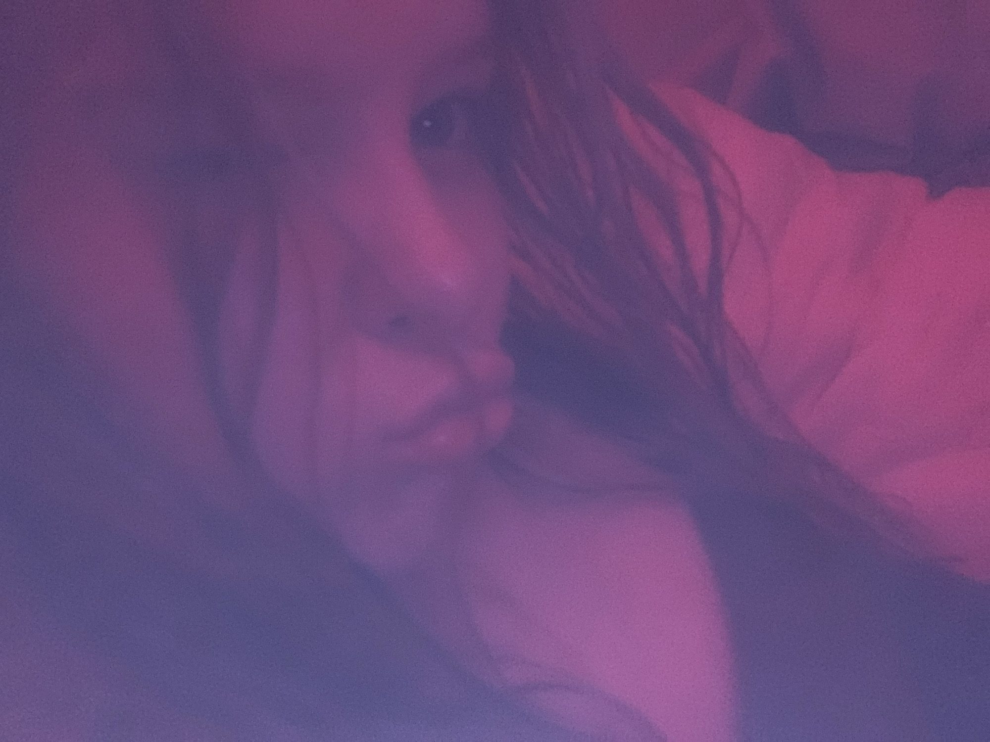 The image presents a heavily filtered and blurry portrait of a person, likely female, captured in a soft, dreamlike aesthetic. The dominant colors are shades of purple, violet, magenta, and pink, creating an atmospheric, low-light setting. The image is significantly grainy, enhancing its artistic and diffused quality.
The primary subject is a person's face, visible on the left side of the frame, angled towards the right. The right eye of the subject is discernible, appearing dark with a small glint of light. Below the eye, parts of the nose and mouth area are softly visible, suggesting a relaxed or slightly parted mouth. Long, dark hair cascades down the right side of the face and extends towards the right edge of the image. The person appears to be lying down or resting, possibly on a soft surface, indicated by the soft forms and what seems to be a light-colored garment or bedding visible on the right.
The activity taking place is passive, suggesting a moment of rest, contemplation, or sleep. The dim, colored lighting implies an indoor setting, likely evening or night, with mood lighting rather than natural daylight. There are no specific location cues beyond an assumed indoor environment. The emotional tone conveyed is calm, serene, and possibly intimate or reflective due to the soft focus and muted expression. There is no visible text in the image. The location context is Unknown, Russia.
