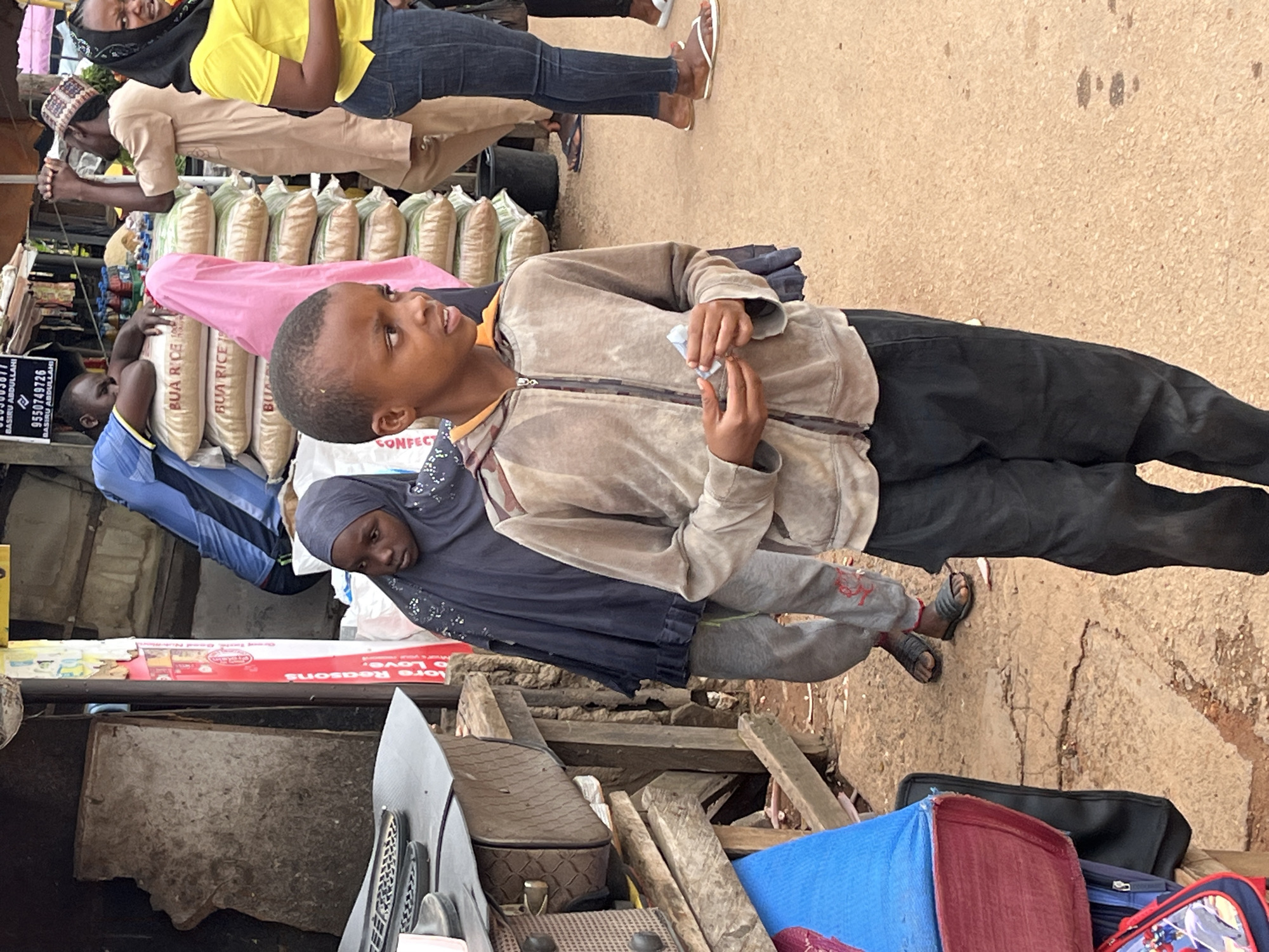 The image depicts a street scene in Jos, Nigeria, possibly a market or a busy roadside. In the foreground, a young boy wearing a light brown hooded jacket and black pants stands looking to his right, holding a small white object in his hands. Behind him and to his left, a young girl wearing a dark blue hijab and gray pants looks to her left. Several large sacks of rice are stacked behind the children, with text that reads "BUA RICE". Other individuals are visible in the background, including a person in a blue shirt, a person in a tan outfit with a patterned cap, and a woman in a yellow shirt. The environment appears to be an outdoor setting with a dirt or gravel ground, and some wooden structures and stalls are present. The overall scene suggests a typical day in a bustling urban environment in Nigeria.