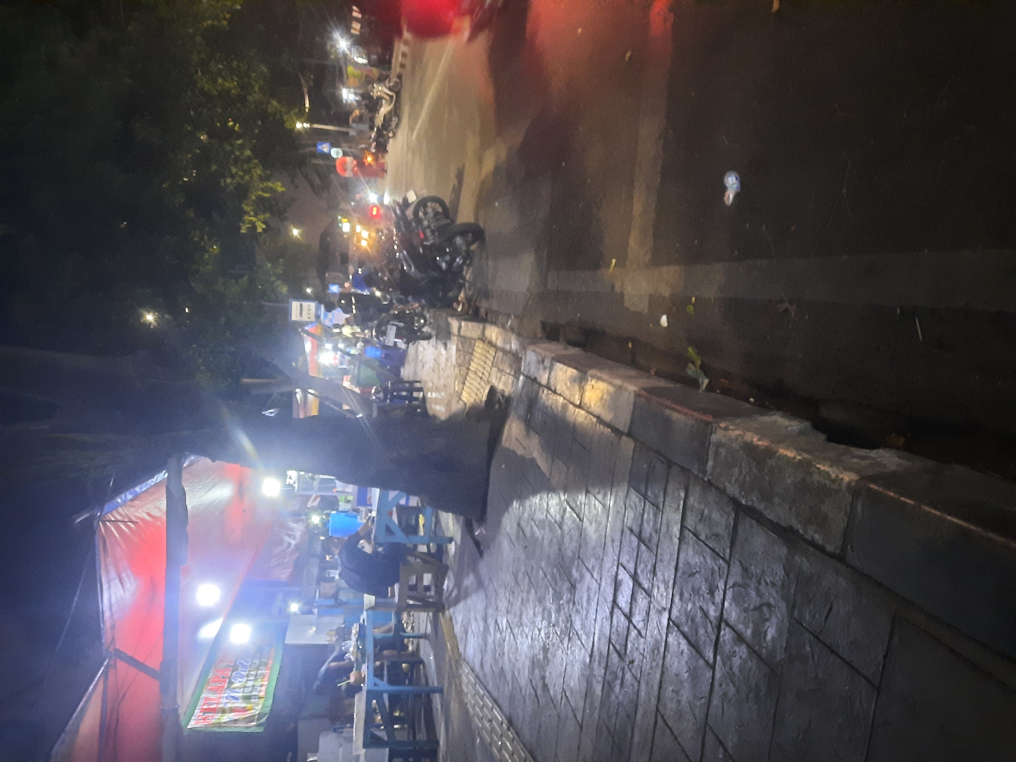 A nighttime street scene in North Jakarta, Indonesia, depicts a row of outdoor food stalls situated along a sidewalk. Red and white tarps cover the stalls, illuminated by bright lights, casting a warm glow. Several tables and chairs are set up on the paved sidewalk, some occupied by patrons. A large tree trunk is prominent in the foreground, casting a shadow across the walkway. Beyond the stalls, motorcycles are parked and some are in motion on the street, with their headlights and taillights visible. The street appears wet, possibly from recent rain, reflecting the lights. The overall atmosphere suggests a busy, local gathering spot for food and social interaction.