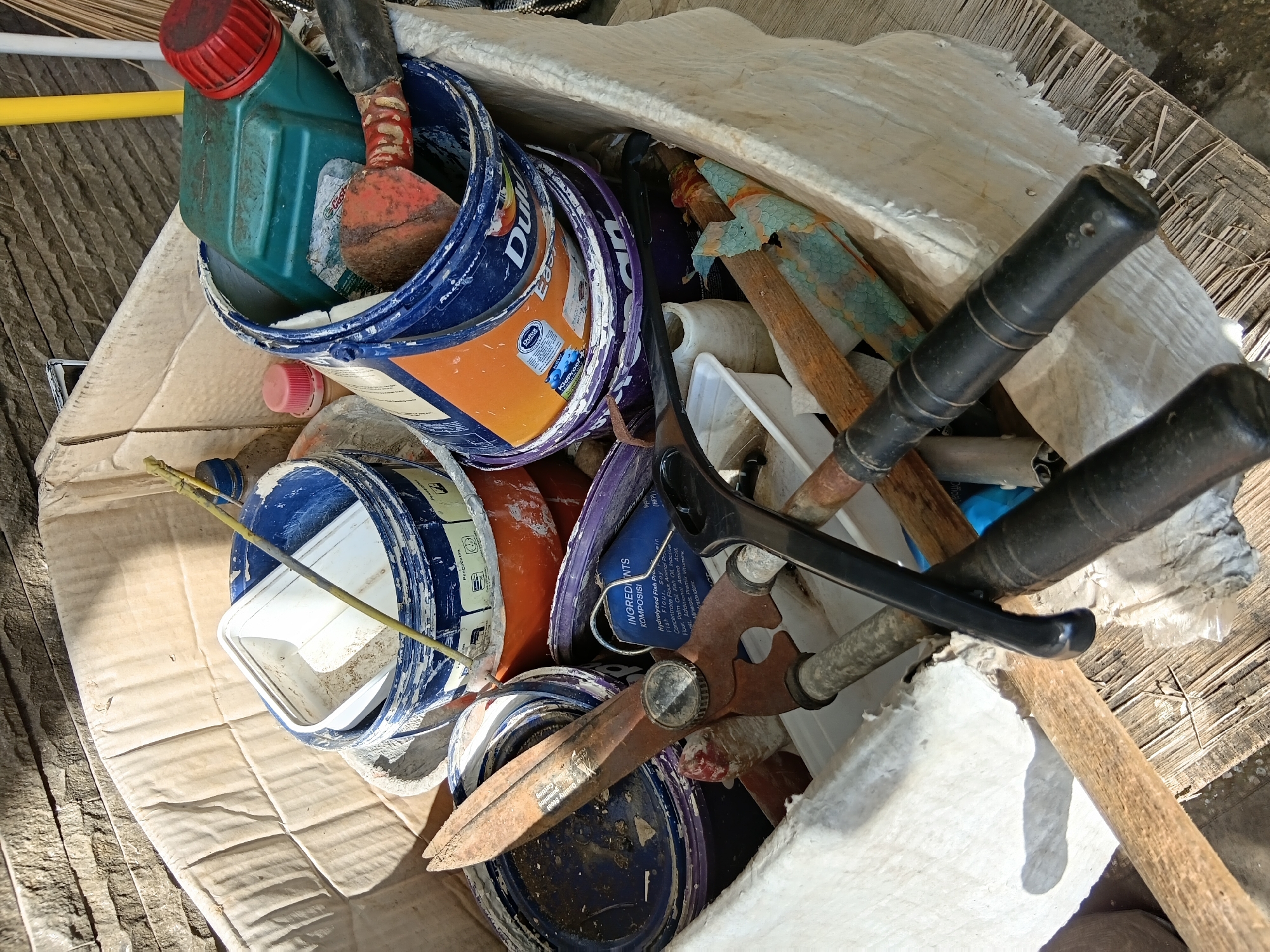 A cardboard box is filled with various used items, primarily paint cans and gardening tools, placed outdoors in the city of Surabaya, Indonesia. The box appears weathered and is situated on a surface composed of rough wooden planks. Sunlight casts shadows on the objects within the box, suggesting it is daytime. Among the visible items are several plastic paint cans, some with their lids on, others open and containing residue. A green jerrycan with a red cap is also present. Gardening tools include a pair of large, rusty shears, a black-handled tool that resembles wire cutters, and wooden-handled implements. The background shows a stone wall with a rough texture. The overall scene suggests a collection of discarded materials, possibly awaiting disposal or repurposing. There are no people visible in the image, and no actions are taking place other than the static placement of the objects. The environment is outdoors, with indications of a somewhat unkempt area.