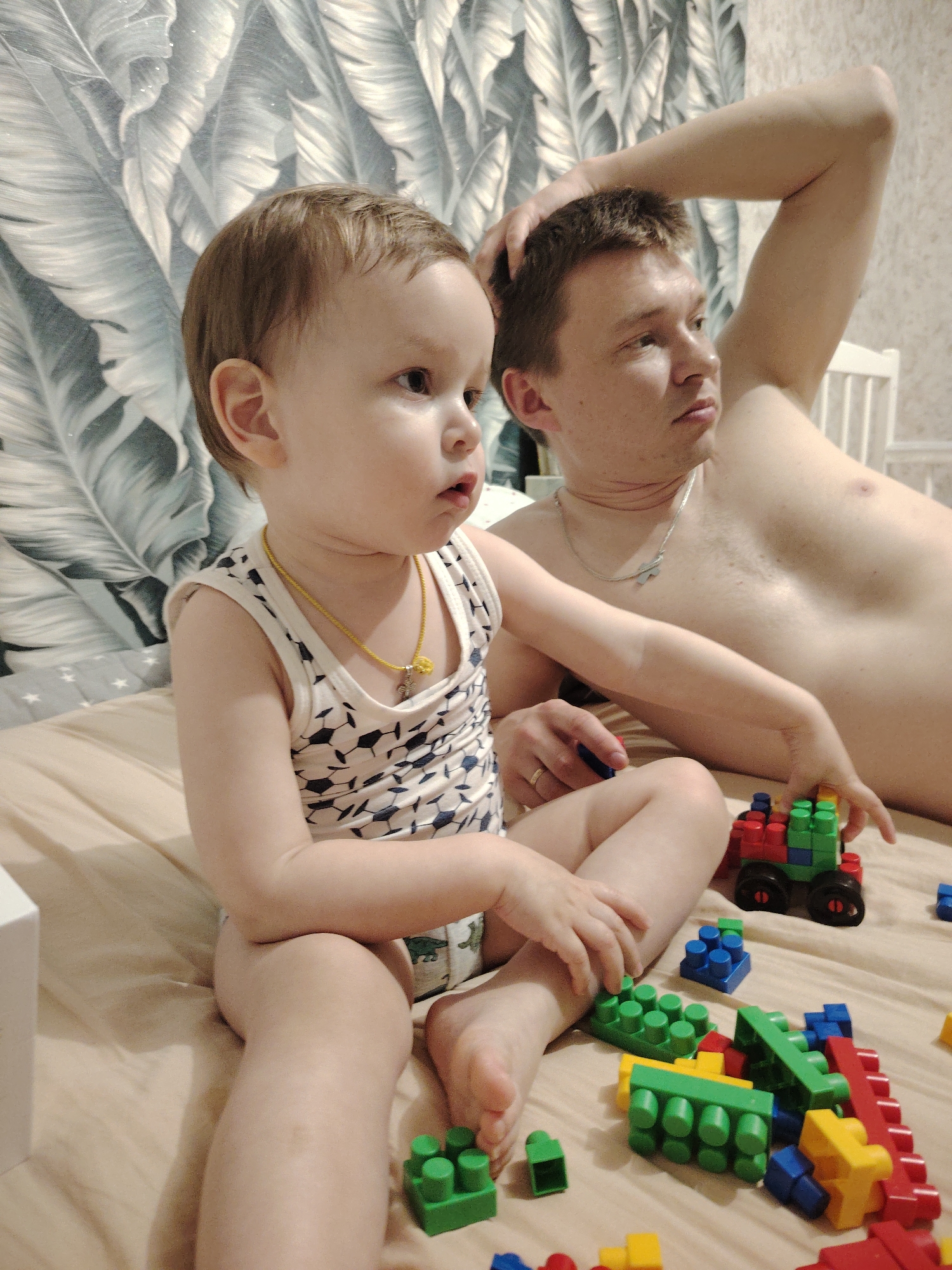 A low-angle, indoor shot shows a fair-skinned toddler in the foreground and a fair-skinned adult male in the background, both on a bed. The toddler, likely a young child around 1-2 years old, is sitting cross-legged, wearing a white tank top with a geometric soccer ball pattern and a gold necklace. They appear to be looking to the left. Around them are several brightly colored plastic toy blocks and a small toy vehicle.
The adult male is lying on his back with his right arm raised behind his head. He is shirtless, wearing a silver necklace and ring. He is looking in the same direction as the toddler. The setting appears to be a bedroom, with a wall covered in a leaf-patterned wallpaper visible behind them. The bed is covered with a plain beige sheet.