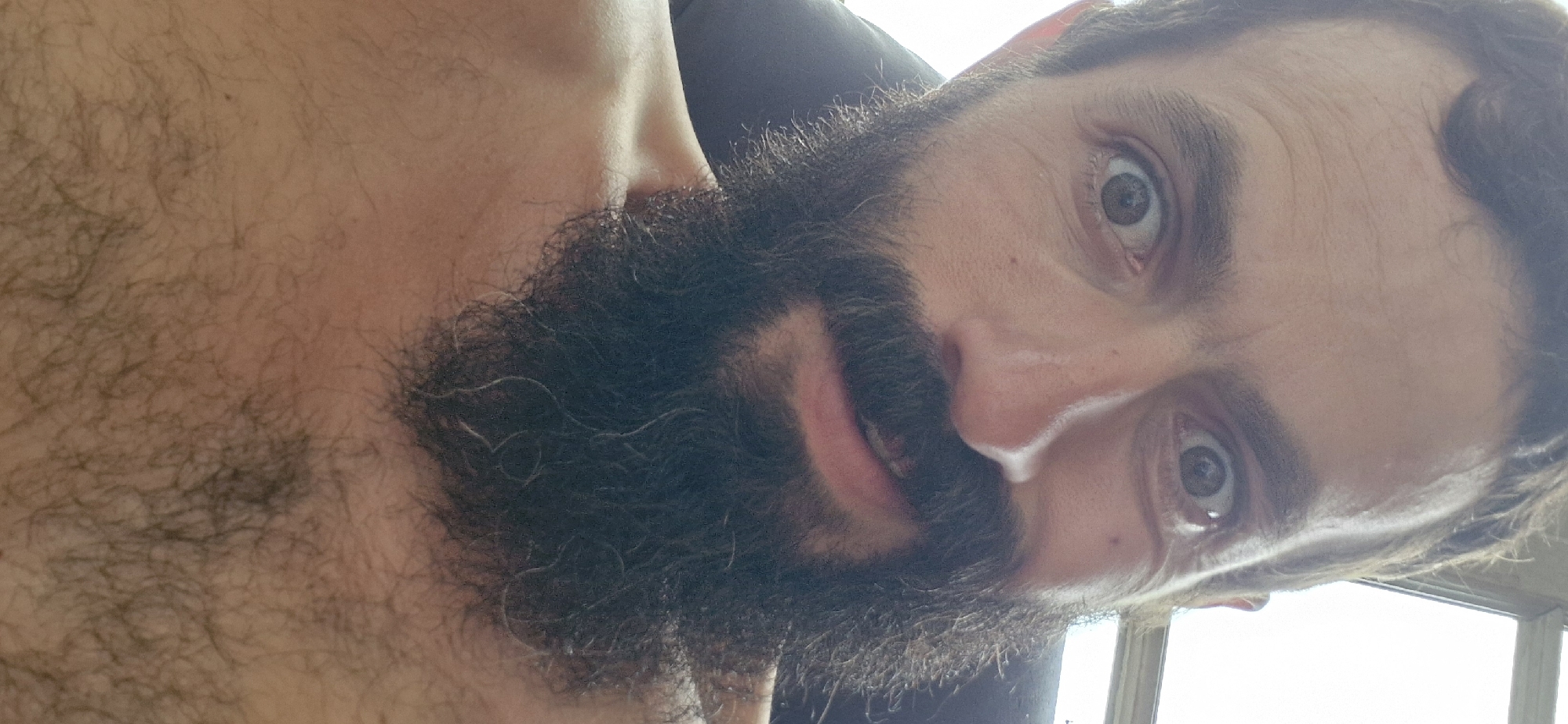 The image is a close-up, selfie-style portrait of a man with a dark beard and mustache, cropped from the chest up. He has wide, expressive eyes with visible wrinkles around them, and his mouth is slightly open, revealing his teeth. His hair is dark and appears somewhat unkempt. The background is out of focus, but suggests an indoor setting with a window or light source to the right. The man is shirtless, and his chest and upper abdomen are visible, covered in dark hair. The scene appears to be taken in a casual, personal environment.