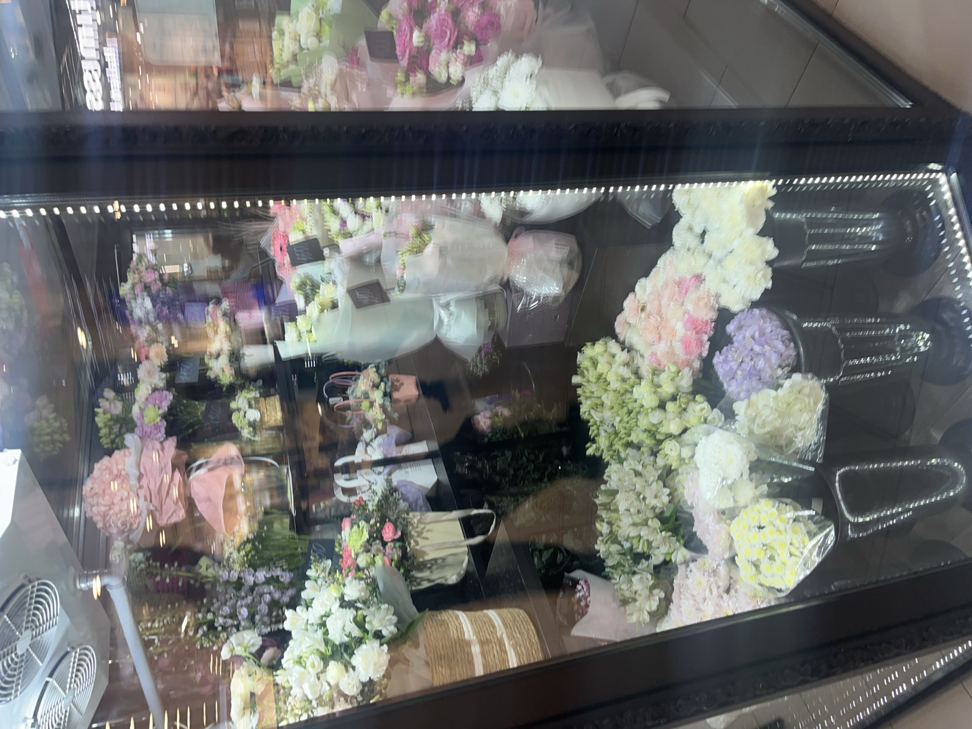 The image displays a tall, glass-fronted refrigerated display case filled with a vibrant assortment of fresh flowers, likely situated within a flower shop in Ulyanovsk, Russia. The display case features a dark frame and is illuminated internally by vertical LED strip lights along its left and right edges.
Inside, numerous bouquets are arranged upright, showcasing a wide variety of flowers in shades of pink, purple, white, cream, light green, and yellow. Common types appear to include hydrangeas, carnations, roses, and various filler flowers. Many bouquets are individually wrapped in clear plastic, delicate pink paper, or other light-colored materials, suggesting they are prepared for individual sale. Some arrangements are presented in small woven baskets or gift bags, adding to their appeal. Small, dark tags are attached to several bouquets, but the text on them is illegible.
On the bottom-left exterior of the display case, a metal fan or cooling unit with two circular grilles is partially visible, indicating the refrigeration system designed to keep the flowers fresh. The glass front of the case reflects the surrounding interior environment, showing blurred lights and indistinct details. Notably, a reflection in the upper-right section of the glass clearly shows the Russian word "КРАСИВЫЕ" (Krasivyye), meaning "beautiful," which serves as a location cue consistent with the provided context of Ulyanovsk, Russia.
The scene depicts a commercial retail setting where flowers are artfully presented for customers to browse and purchase. No people are visible, and the time of day or weather cannot be determined from the indoor setting.