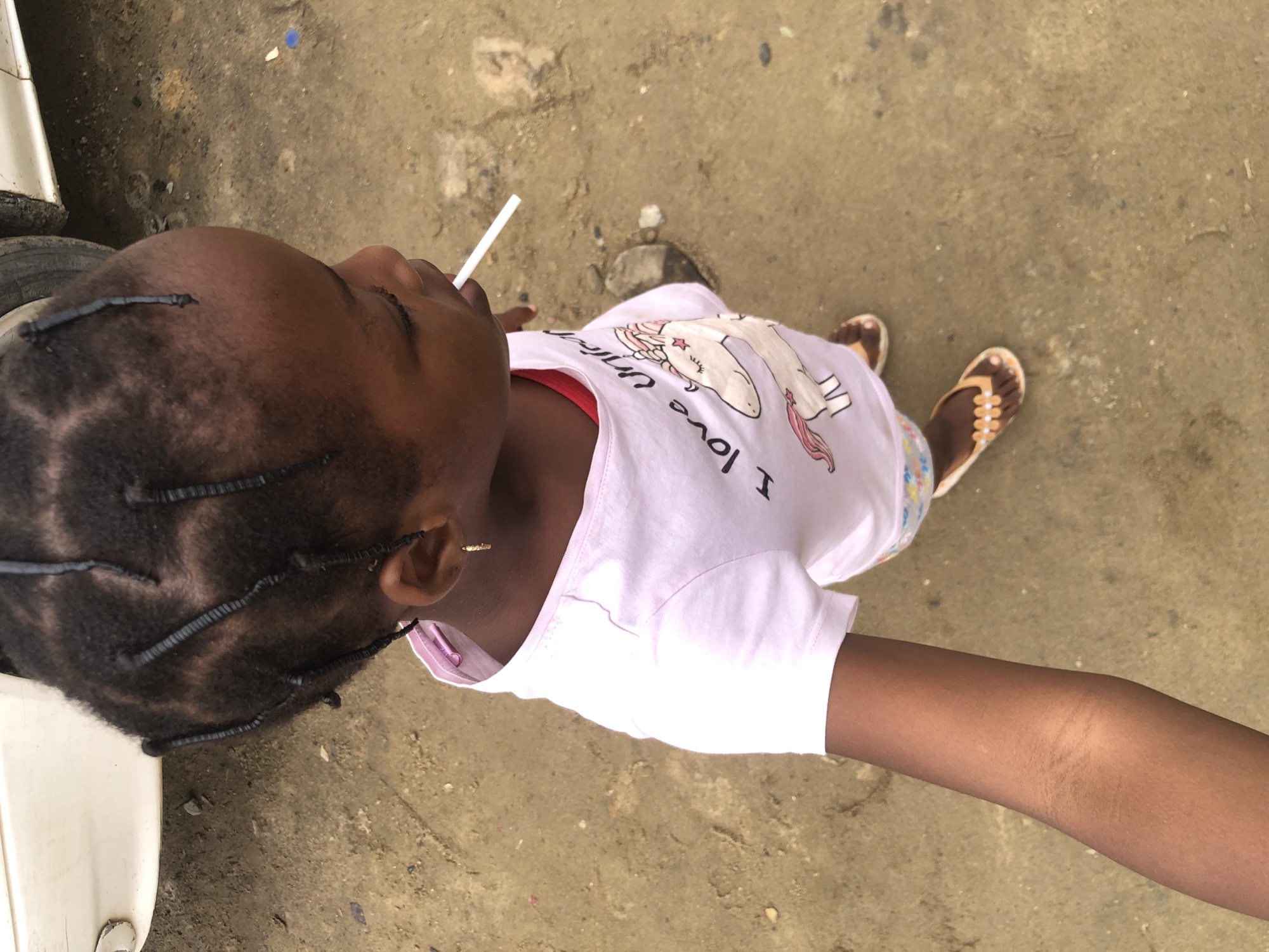 A young Black child, appearing to be between the ages of 3 and 5, is the sole subject of this overhead photograph taken outdoors, likely on a dirt or unpaved surface in Ado, Nigeria. The child is facing to the right and slightly downwards, with their left arm extended towards the camera. They have dark skin and dark hair styled in numerous thin braids, some of which are visible on top and at the back of their head. A small, gold hoop earring is worn in their left ear.
The child is wearing a light pink t-shirt with a graphic print of a unicorn and the text "I love unicorn." Beneath the t-shirt, a red garment is visible. The child's legs are covered by patterned shorts, only partially visible. On their feet, they wear brown and orange flip-flops. In their mouth, the child has a white lollipop, with the stick protruding to the right.
The environment appears to be a simple, unkempt outdoor setting. The ground is a muted brown, with scattered pebbles and debris. In the upper left corner of the image, a portion of a white vehicle's body and a tire are visible. The lighting suggests it is daytime, with some shadows present, indicating a sunny or partly cloudy sky. The overall impression is a candid, informal snapshot of a child in a casual outdoor environment.