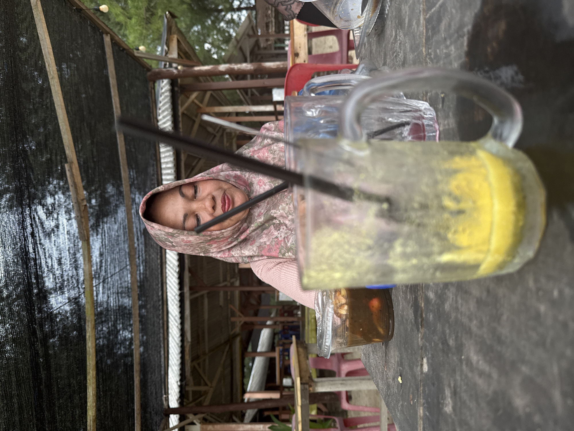 In Meunasahbale, Indonesia, a woman wearing a pink and green floral hijab and a pink top sits at a weathered wooden table outdoors. She is looking down at a small clear plastic container filled with a dark, chunky food item and possibly some red elements. In front of her, a large glass mug with a handle contains a yellow liquid with remnants on the side, and a black straw. Another glass, mostly out of focus, is visible to the right of the first. In the background, a rustic open-air structure with a corrugated roof and wooden supports is visible, suggesting a casual dining or seating area. The overall setting appears to be a food stall or outdoor restaurant, likely near a beach or coastal area, given the presence of a black mesh canopy overhead and vegetation in the distance.