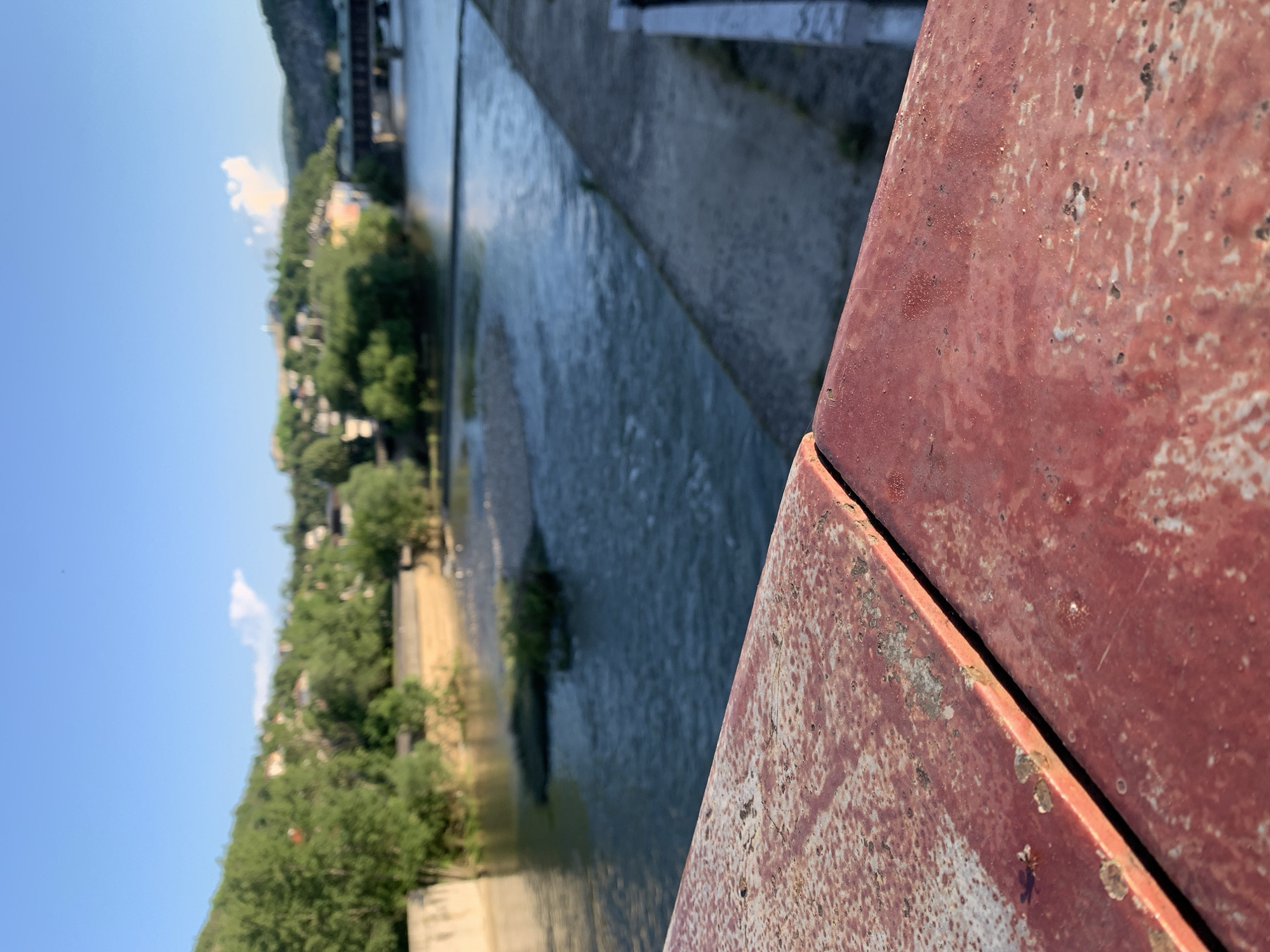 A tilted, slightly blurred, high-angle shot shows a water channel with a city scene on one side, taken from the perspective of a rusty metal structure. The channel runs horizontally across the image. The city, featuring green trees and buildings, is to the left of the channel, with a blue sky above. The right side of the image is dominated by the reddish-brown rusty metal, with a clearly defined edge indicating the structure's shape. The location of the city is unknown.