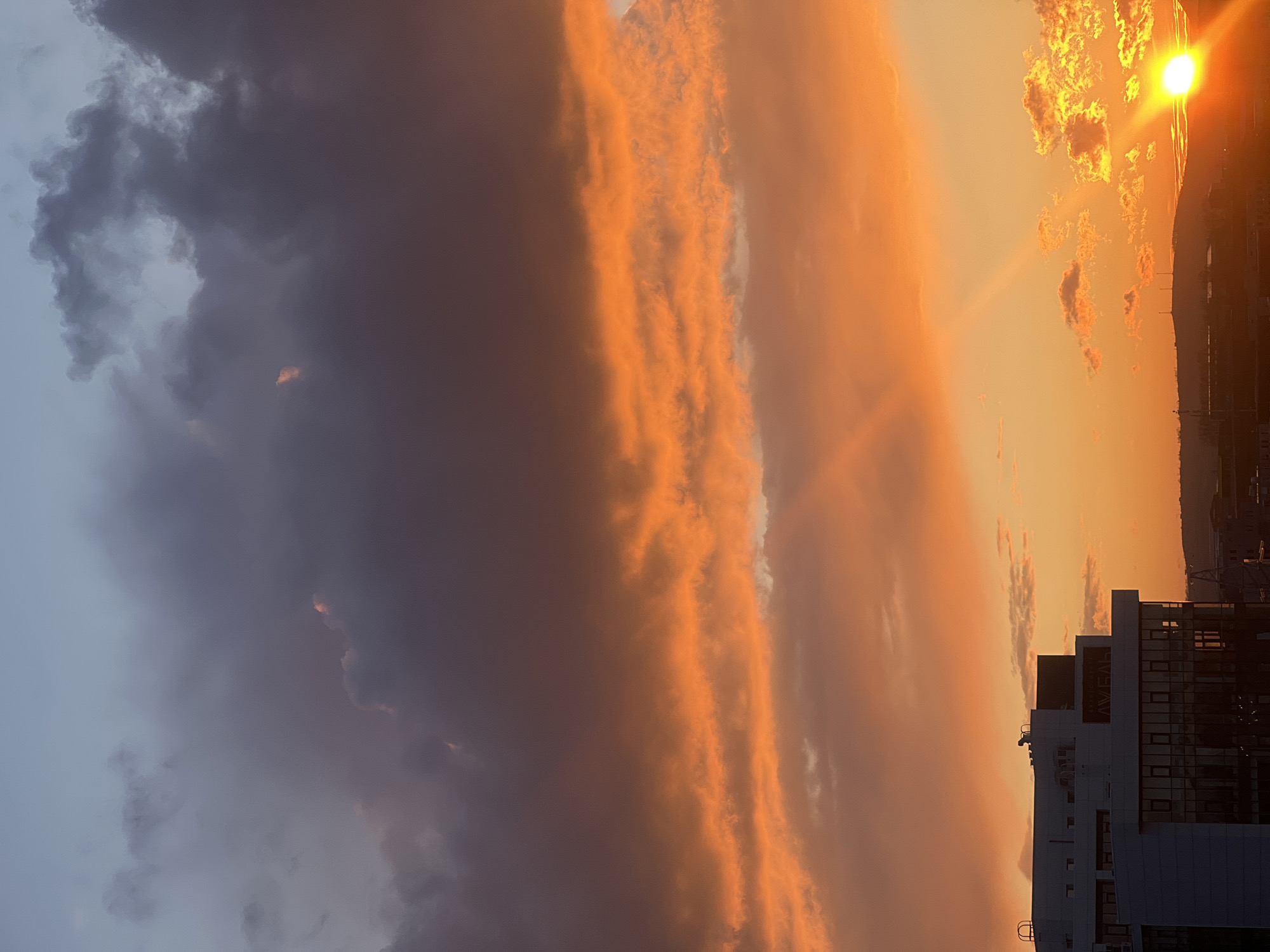 The image shows a sunset view in Krasnoyarsk, Russia. The sky is filled with clouds of varying colors, ranging from gray to orange. The sun is visible on the right side of the image, partially obscured by clouds. A modern building is silhouetted in the foreground against the sky, with a sign that says "AVEAH" in Russian. In the background, a cityscape is vaguely visible on the horizon.