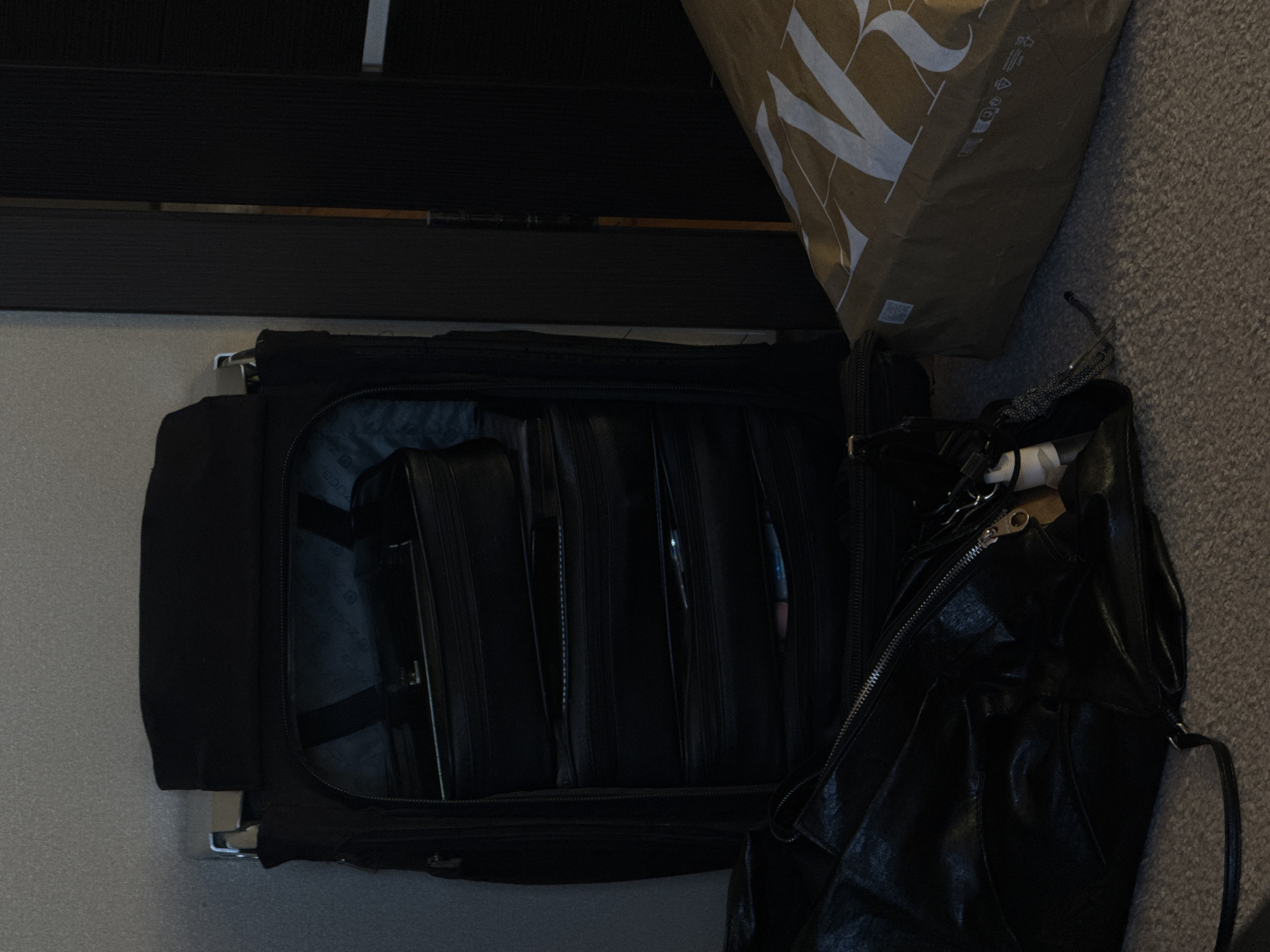 A black suitcase filled with black briefcases is open on the floor, next to a Zara shopping bag and a black purse. The location appears to be indoors.