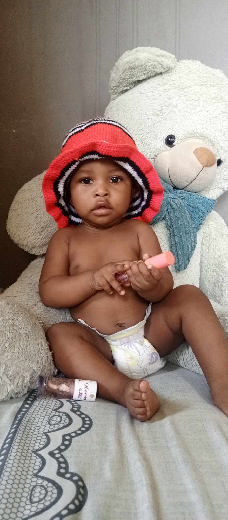 An indoor, vertical shot features a baby sitting in front of a large teddy bear. The infant, with dark skin, wears a colorful knitted bucket hat with red, black, and white stripes, and a light-colored diaper with subtle patterns. The baby is shirtless and looks directly at the camera with a neutral expression. In its hands, the baby holds two small objects: a pink cylindrical item and a smaller, clear object.
Behind and to the right of the baby is a large, light-colored (possibly off-white or light grey) fuzzy teddy bear with black button eyes and a light blue fabric tied around its neck. The teddy bear appears to be providing support or simply part of the setup.
The baby and teddy bear are seated on a bed or soft surface covered with a light-colored sheet featuring a grey, ornate, lace-like pattern along its edge. To the left of the baby's leg, a small, clear bottle with a label is lying horizontally on the sheet. The label prominently displays the text "Honey Mimi" and includes a small graphic resembling a diamond or crown.
The background consists of a plain, light brown or grey wall with faint vertical lines, suggesting a simple domestic setting. The overall mood of the image is calm and innocent, capturing a moment with the baby.