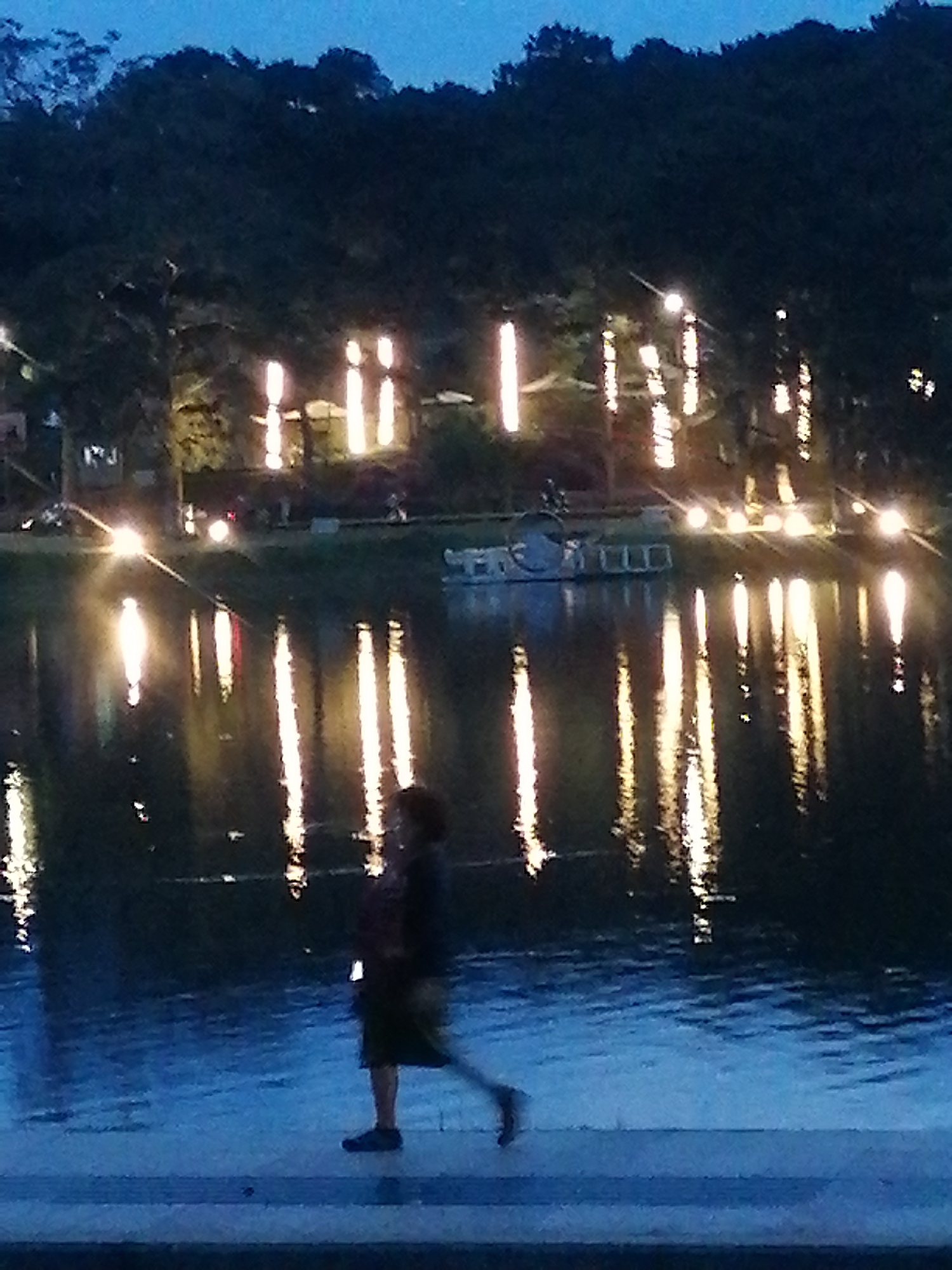 The image depicts a nighttime scene at Xuan Huong Lake in Da Lat, Vietnam. The lake's surface reflects the lights from trees and structures along its edge, creating streaks of illumination. In the foreground, a person is walking along a paved pathway adjacent to the lake. The person is silhouetted against the water, wearing dark clothing and a backpack, and appears to be in motion. The background consists of a dark treeline illuminated by numerous vertical lights, possibly decorative string lights or lanterns, casting a warm glow. The overall atmosphere is serene and slightly blurred due to the low light conditions.
