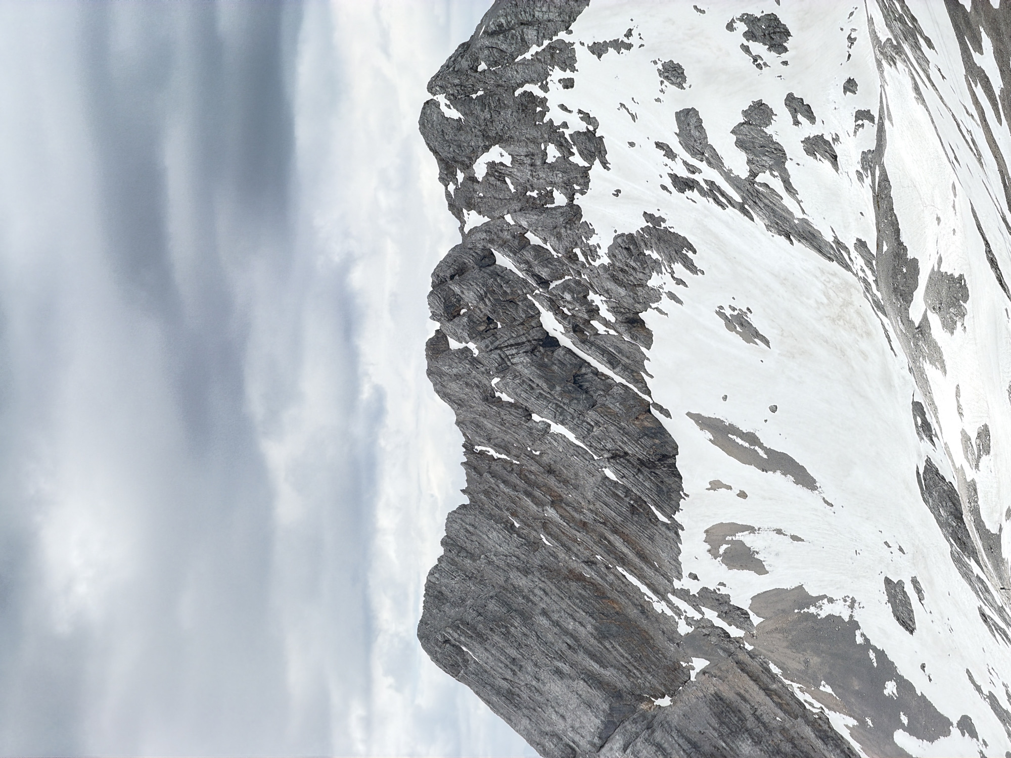 The media file shows a close-up, vertical view of a snow-covered mountain face with rock formations. The mountain is rugged, with streaks of gray rock contrasting against patches of white snow. The snow appears unevenly distributed, suggesting a steep and rocky terrain. The lighting indicates it is daytime, and the overcast sky, characterized by soft, diffused light and cloudy textures, suggests a cool, possibly windy, mountain environment.
The scale of the mountain is implied by the fine texture of the rock and snow, hinting at a vast, natural landscape. The image captures the raw, untamed beauty of the alpine environment. There are no visible people or man-made objects, emphasizing the natural setting. The overall mood is one of serene, grand natural beauty, highlighting the power and scale of the mountain. The location context provided is Garmisch-Partenkirchen, Germany, which is known for its high alpine scenery.