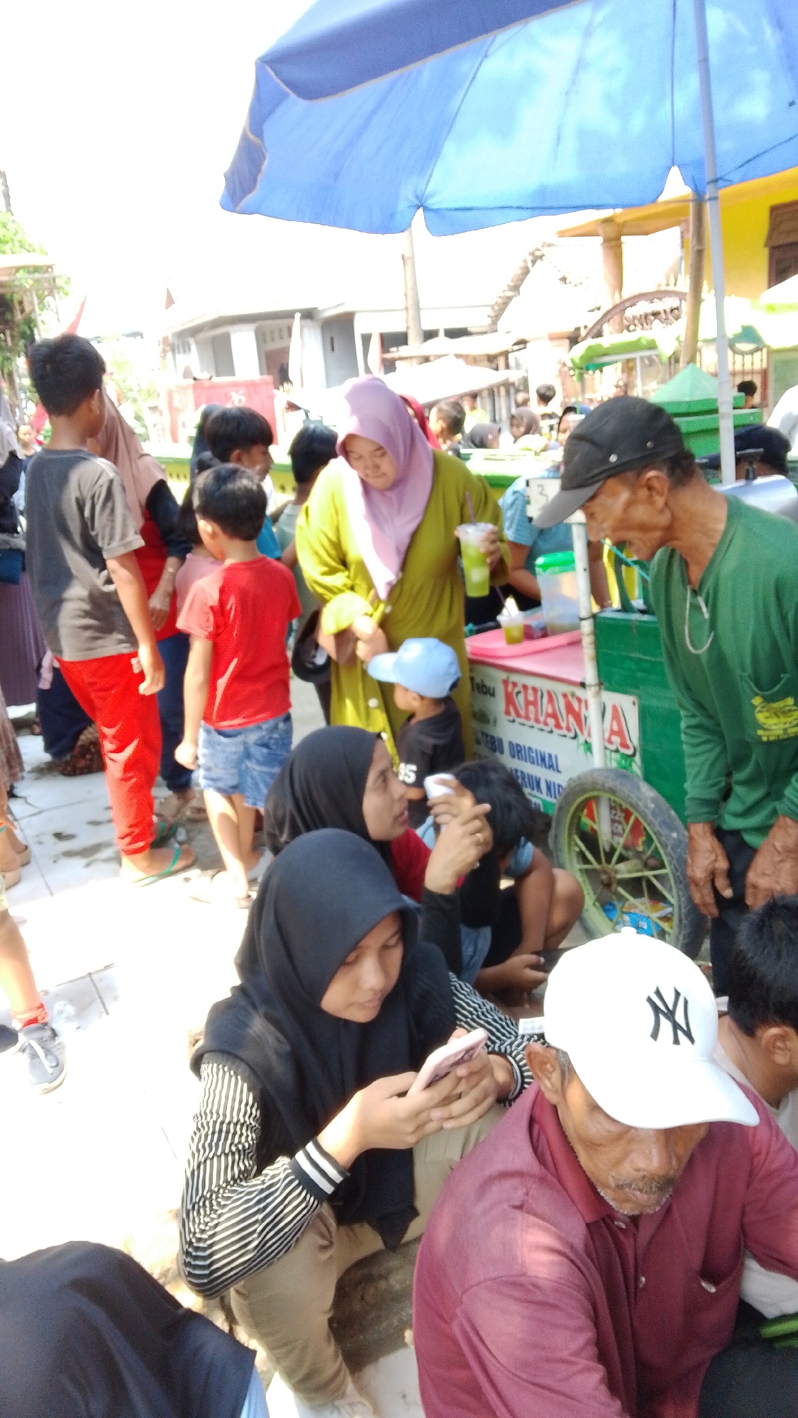In Mancar, Indonesia, a group of people are gathered outdoors under a large blue umbrella, likely at a street-side stall. The focus is on a vendor with a green cart selling drinks. An older man wearing a green shirt and a black cap is speaking to someone. A woman in a lime green hijab and dress holds a beverage. Nearby, a young boy in a blue cap looks towards the vendor. In the foreground, a young woman in a black hijab with a striped shirt is looking at her phone. Another person in a maroon shirt and a white baseball cap with a black logo is seated. The ground appears to be paved or dirt. The background shows buildings and other stalls. The overall atmosphere suggests a busy market or gathering.