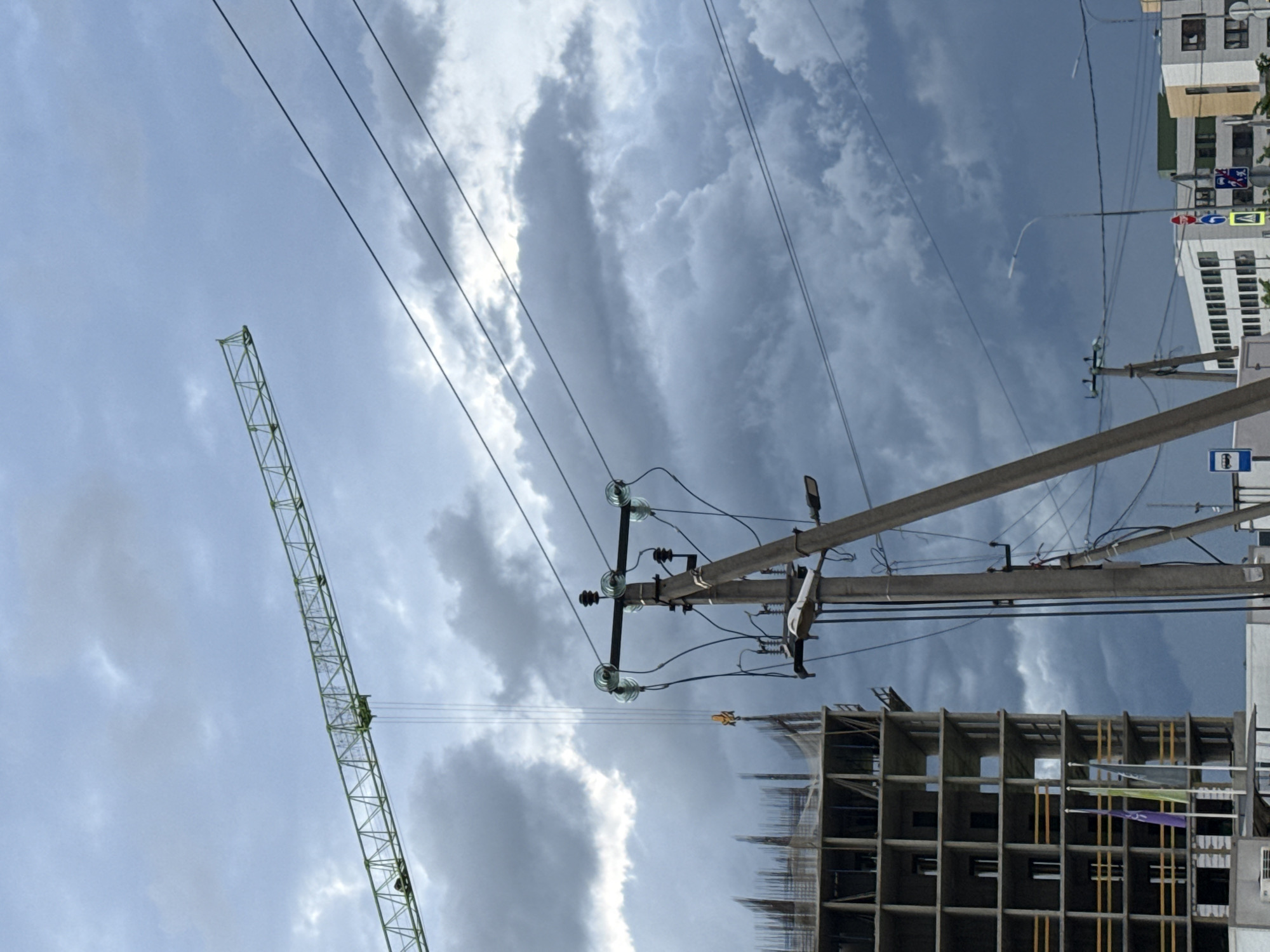 The image depicts a construction site located in a city of Unknown, Unknown. A green crane stands tall against a cloudy sky. In the foreground, there's a building under construction, with its concrete framework visible. Power lines stretch across the sky, supported by utility poles. A street and buildings with traffic signs are visible in the background.