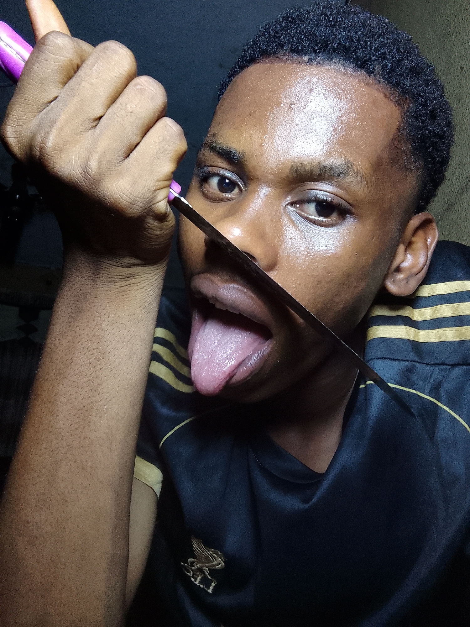 The image shows a young, dark-skinned person with short, dark hair holding a knife horizontally in front of their face, with the blade appearing to bisect the open mouth and extended tongue. The person is wearing a black and yellow striped shirt. The background is dark and indistinct. The location of the scene is not discernible from the image.