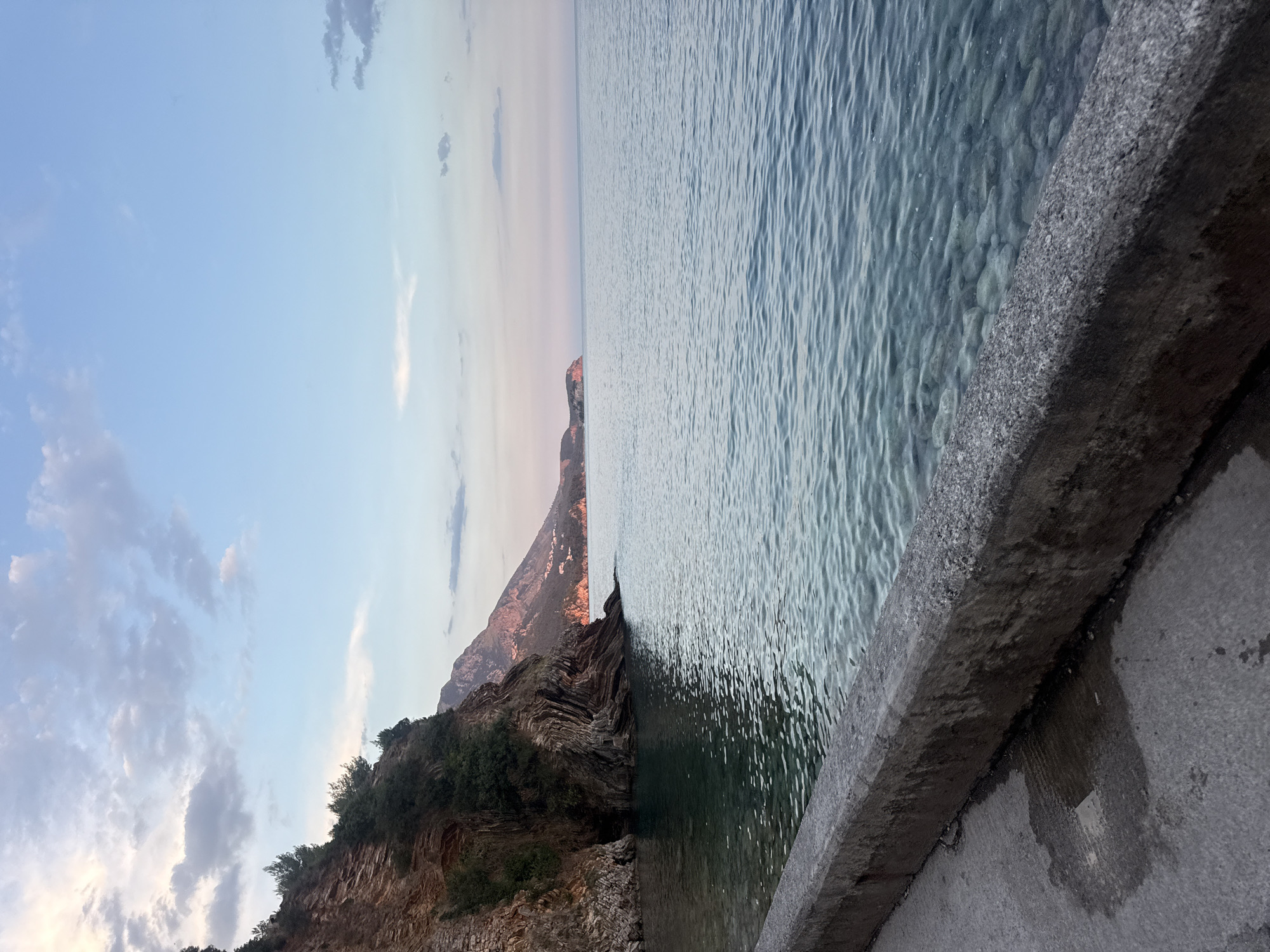 This is a scenic photograph taken from a concrete walkway or pier overlooking the Adriatic Sea in Rafailovići, Montenegro. The water is clear and shallow, revealing smooth, rounded pebbles on the seabed. To the left, a steep, rocky cliff face rises, dotted with sparse green vegetation. The cliff exhibits distinct layers and textures, with evidence of erosion and stratification. In the distance, a rugged coastline stretches out, with mountains in the background. The sky is a soft blend of blue and pastel hues, with wispy clouds, suggesting either dawn or dusk. The overall atmosphere is tranquil and serene. There are no people or visible actions depicted in the scene.