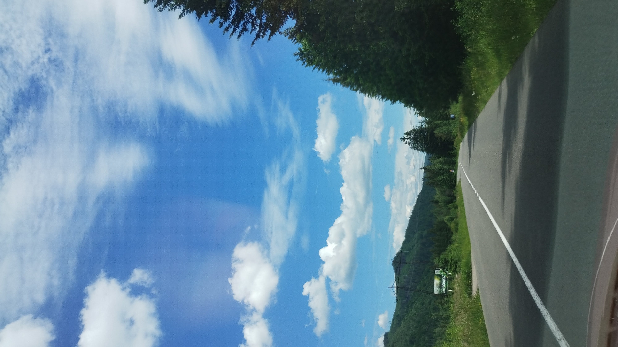 The image depicts a rural road winding through a forested mountainous landscape in Myslivka, Ukraine. The road is paved with a white line marking its center. Lush green trees, including tall conifers, line the right side of the road and cover the hills to the left. The sky is a vibrant blue with scattered white clouds, some appearing wispy. Power lines and pylons are visible on the left hillside. A green sign with white text is also present on the left side of the road, suggesting a location or informational marker within Myslivka. The perspective suggests the image was taken from a vehicle moving along the road.