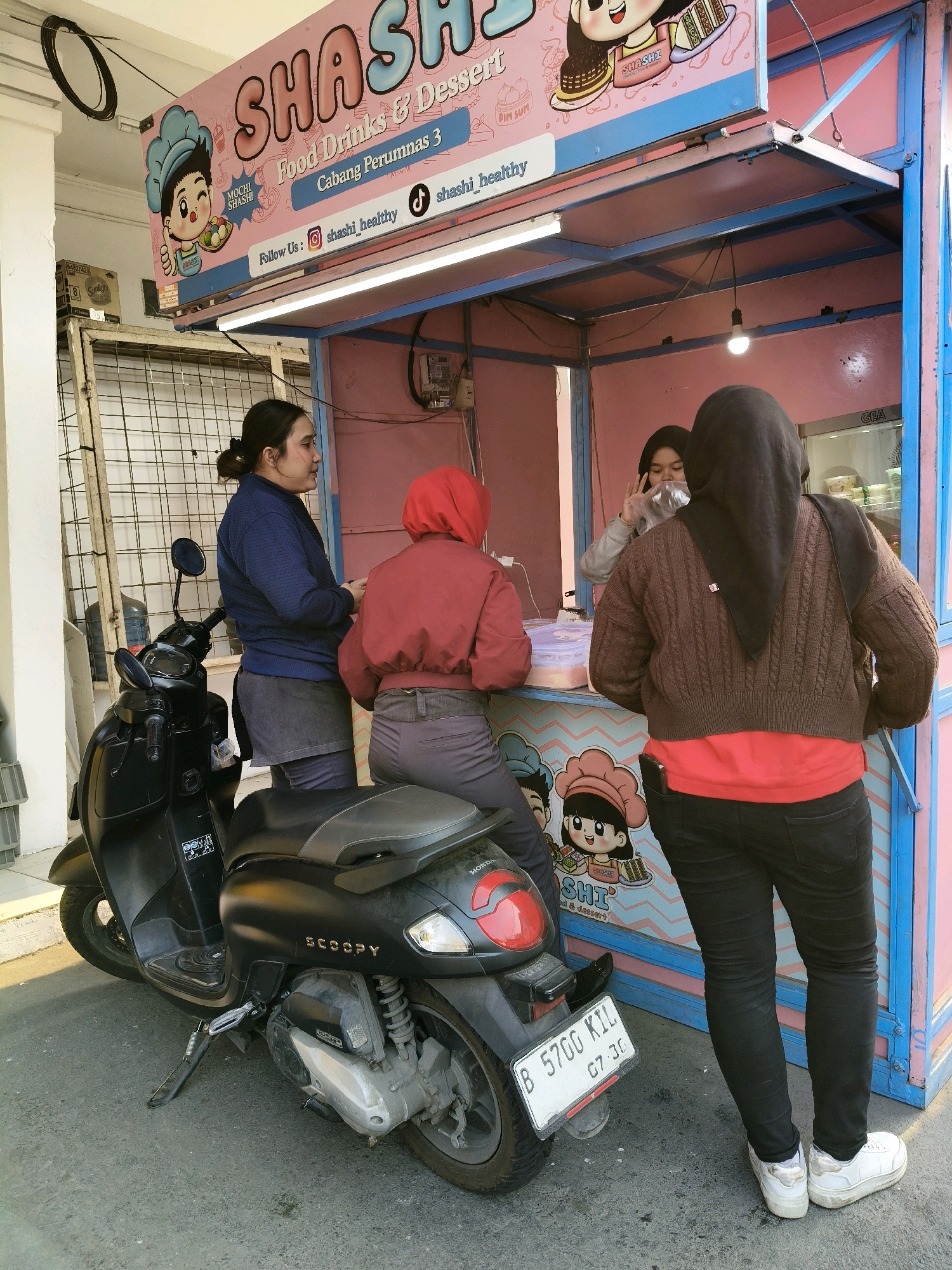 In Bekasi, Indonesia, a street food stall named "SHASHI" displays "Food Drinks & Dessert" and "Cabang Perumnas 3". A black Honda Scoopy motorcycle is parked to the left of the stall. Three individuals are present. One woman, wearing a blue jacket and a dark skirt, stands near the motorcycle. Two other women are at the counter of the stall. One, wearing a red hooded jacket and grey pants, is facing away from the camera. The second woman, wearing a brown knitted sweater and black pants, is also facing away and appears to be a customer. A third woman, partially visible behind a clear plastic bag and wearing a grey top, is serving at the stall. The stall itself is colorful, with a pink and blue theme, and features cartoon characters. The ground is paved and there is a white wall on the left side of the image.