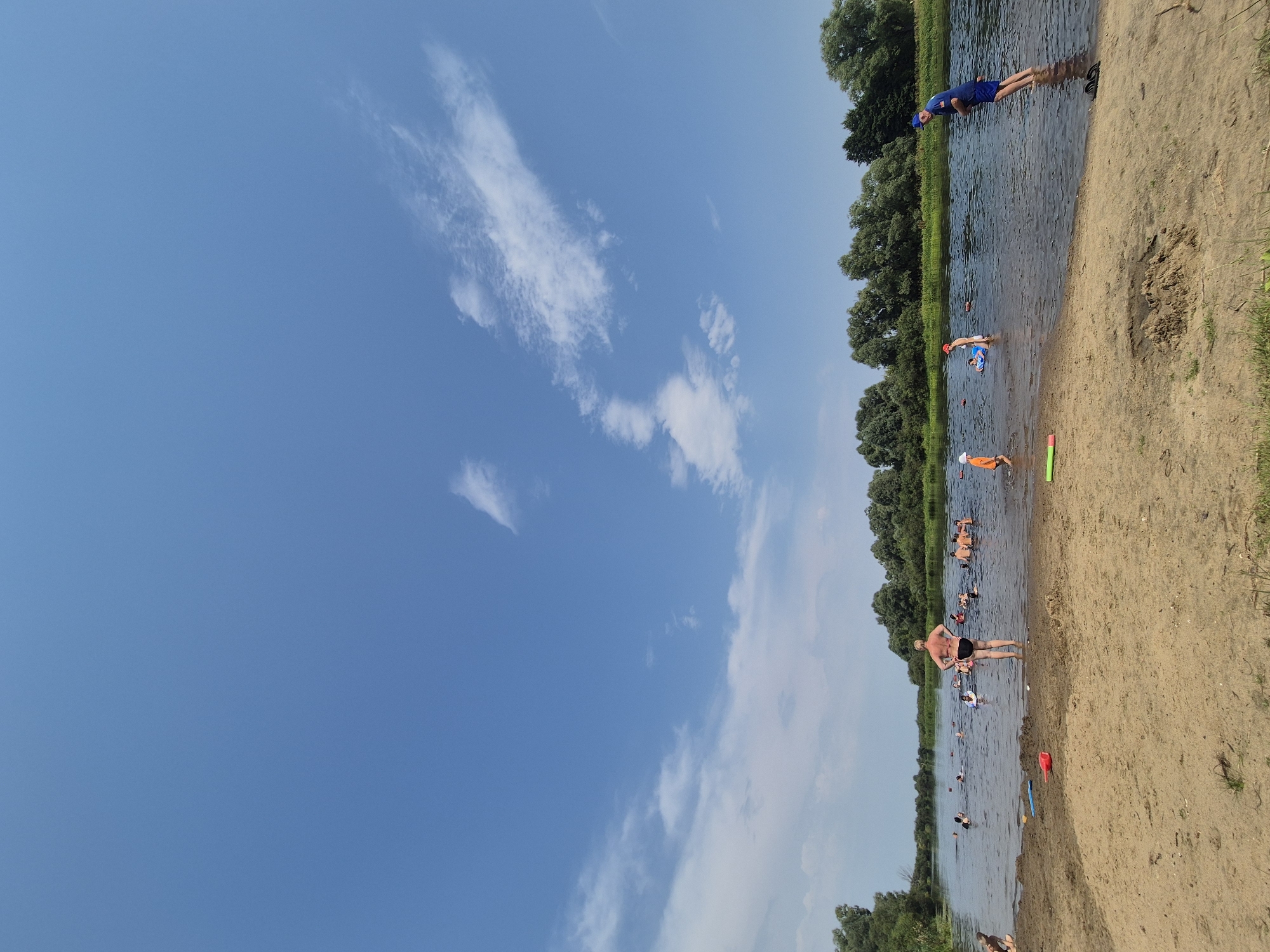 In the city of Parytski Selski Savet, Belarus, a sunny day at a river beach is depicted. The sky is a clear blue with sparse white clouds. A sandy beach occupies the foreground, with scattered blades of grass. The river stretches into the background, teeming with people swimming and enjoying the water. On the beach, a woman in a black swimsuit faces away from the camera, while other individuals are visible in the water and on the shore. In the river, various people are engaged in swimming, some with inflatable rings. The far bank of the river is lined with a dense growth of green trees. Several children are visible in the shallow water, playing. A man in a blue shirt and shorts stands in the water on the right side of the image, facing left. Beach toys, including a red shovel and a green float, are scattered on the sand.