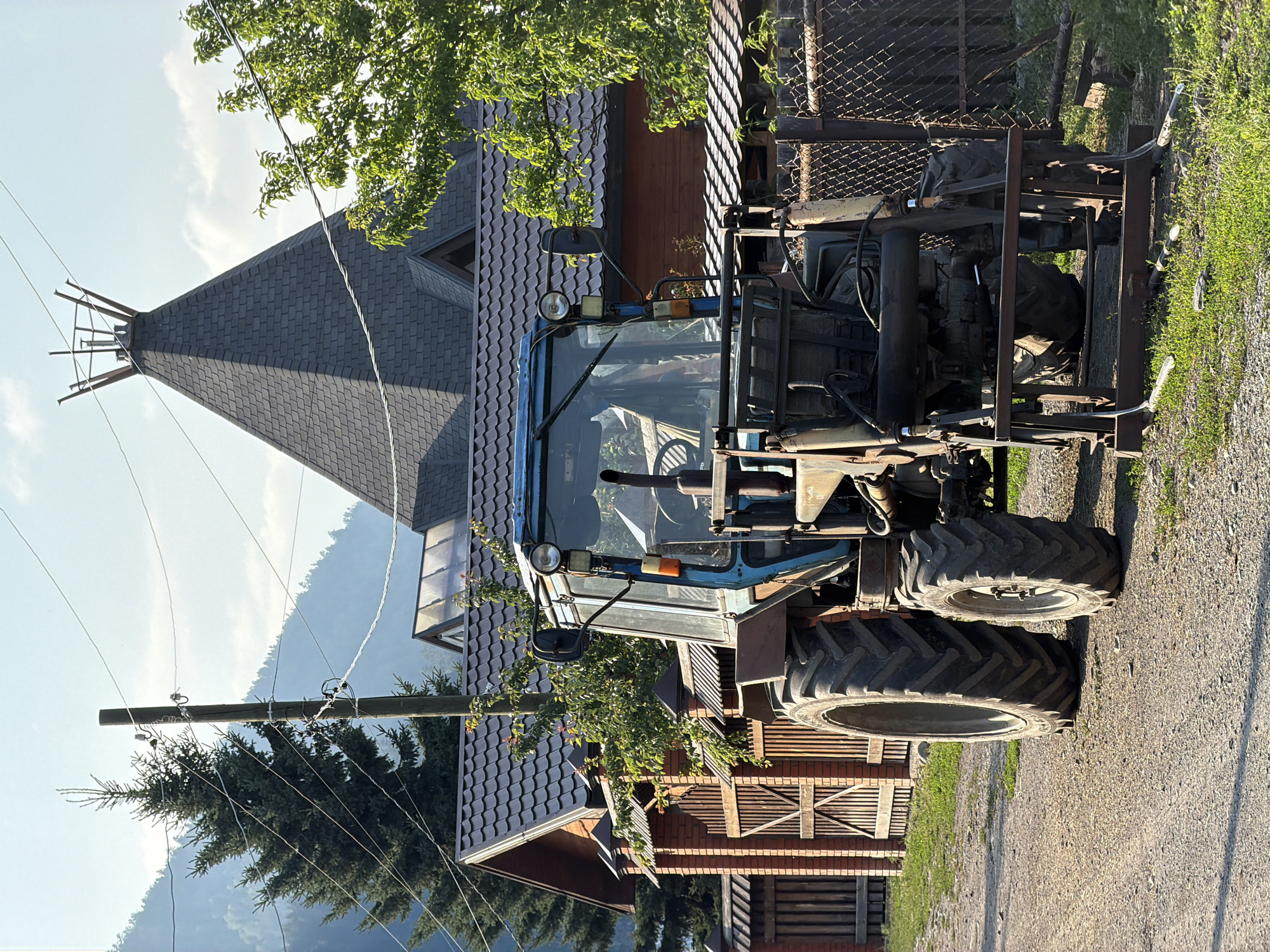 The image depicts an outdoor scene featuring a blue tractor parked on a gravel road in front of a wooden house and fence. The tractor is large and appears to be an older model, with visible wear and tear. It has a front-end loader attachment. Behind the tractor, a traditional-style wooden house with a dark tiled roof is visible, along with a red brick fence. A tall evergreen tree stands to the left of the house, and a lush green tree is to the right. The sky is blue with scattered clouds. The background shows rolling hills or mountains, suggesting a rural or mountainous setting. The overall impression is a rural landscape with agricultural machinery. The image does not contain any people or indicate any specific actions or interactions. The location is identified as Anos, Russia.
