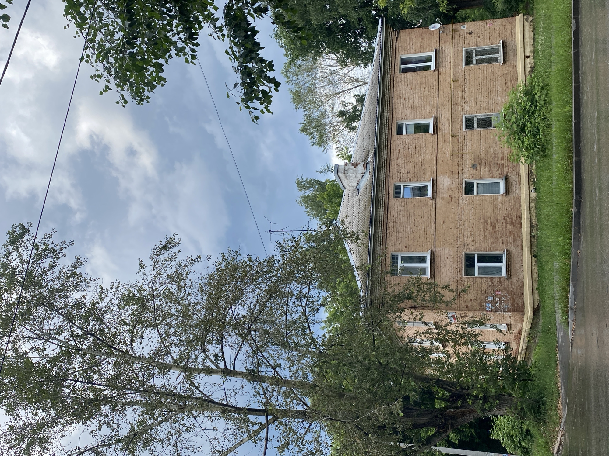 The image depicts a two-story brick apartment building with a grey roof and white-trimmed windows. The building is surrounded by lush green trees and vegetation, with a cloudy sky overhead. There is a large tree with a textured trunk to the left of the building. A paved surface, likely a road or sidewalk, is visible in the foreground, appearing wet and reflective, suggesting recent rain. Electrical wires are strung across the upper portion of the image, and a satellite dish is mounted on the right side of the building. The overall scene is a residential outdoor environment.