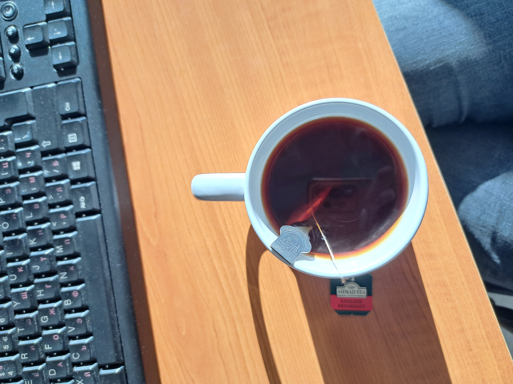 An overhead, close-up shot captures a workspace, featuring a white ceramic mug of tea, a black computer keyboard, and a portion of a person's leg in blue jeans, all resting on a light brown wooden desk.
The central focus is the white mug, filled with dark brown tea. A teabag is actively steeping in the liquid, its string extending to a silver-colored metal tag embossed with a design, perched on the mug's rim. Hanging from the string is a red paper tag that clearly reads "AHMAD TEA LONDON" and "ENGLISH BREAKFAST" in white text.
To the left, a black computer keyboard is partially visible. It features a dual-language layout with both Latin and Cyrillic characters, the latter often appearing in red. This Cyrillic keyboard layout is consistent with its use in Sofia, Bulgaria. The keyboard appears to have some dust accumulation.
In the upper right corner, a segment of a person's leg, clad in blue denim jeans, is visible, suggesting someone is seated at the desk. Strong shadows are cast across the wooden desk surface, indicating a bright light source, likely natural daylight from a window. The overall scene depicts a quiet, personal moment, possibly during work or leisure, with a freshly prepared cup of tea.