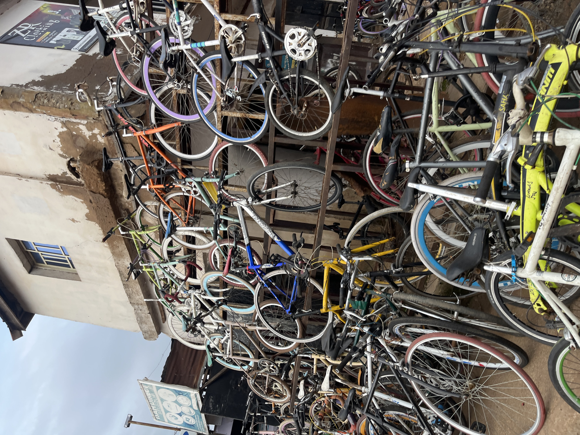 The image shows a densely packed outdoor display of numerous bicycles. The bicycles are of various colors, including white, black, orange, green, purple, and blue, and are leaning against and mounted on metal shelving units. Some bicycles are stacked on top of each other. A building with a textured, light-colored facade and a window with barred panes is visible in the background, as is a sign that reads "DAM DIRECT BELLETTUM". Another sign with "ZR CLOTHING" is also visible. The scene appears to be a bicycle shop or market in Bukuru, Nigeria, indicated by the text on the sign and the general appearance of the urban environment. There are no people visible in the image. The lighting suggests an overcast day.