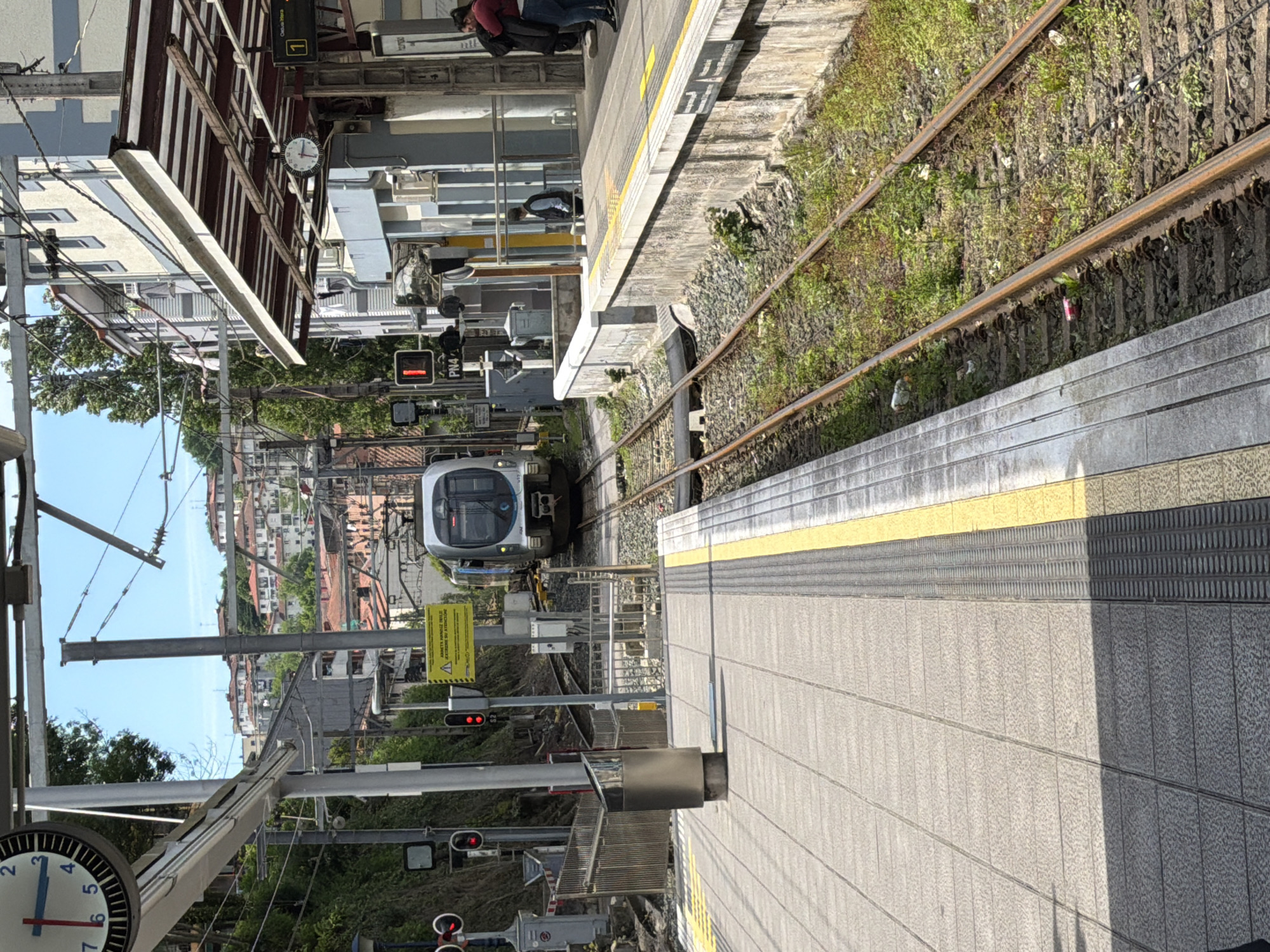 The image depicts a train approaching a platform at a station in Errenteria, Spain. The train is light grey and white, with its front facing the viewer. The platform is grey and divided by a yellow line near the edge. Train tracks run alongside the platform, bordered by gravel and vegetation. Overhead power lines and support structures are visible, along with station signage and signals. A clock is visible on a support beam. The background includes buildings and trees, indicating an urban setting. A few people are visible on a platform further away.