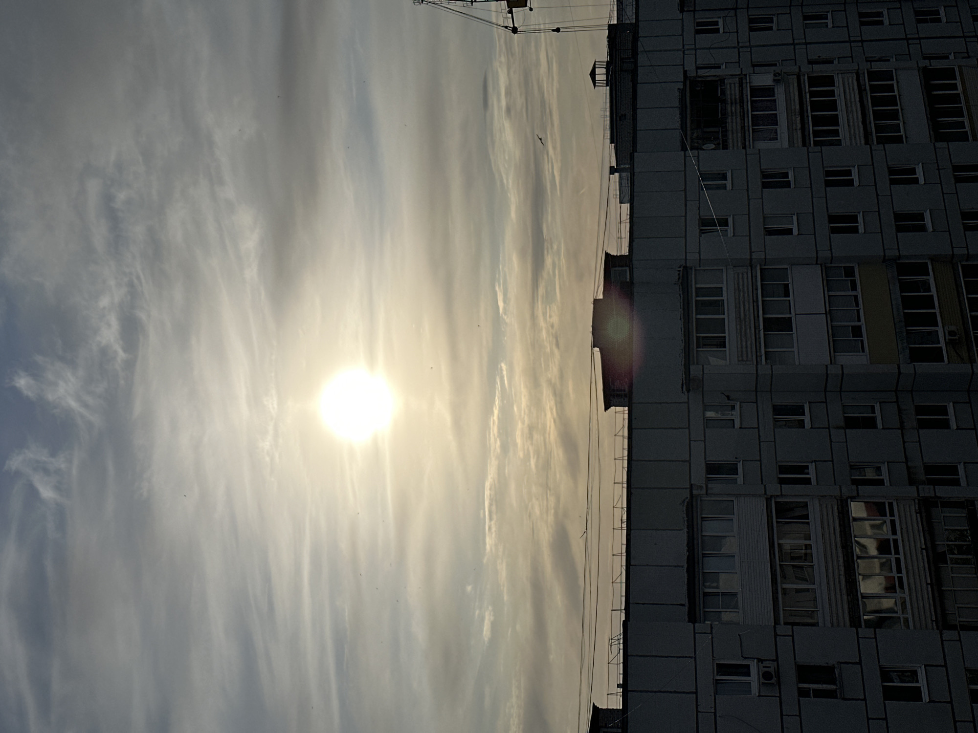 The image shows a tall apartment building in Chaikovsky, Russia, under a bright, cloudy sky. The sun is visible, shining through the clouds. The building is a Soviet-era construction with a repetitive pattern of windows and balconies. There are signs of a crane or other construction equipment on the top of the building.