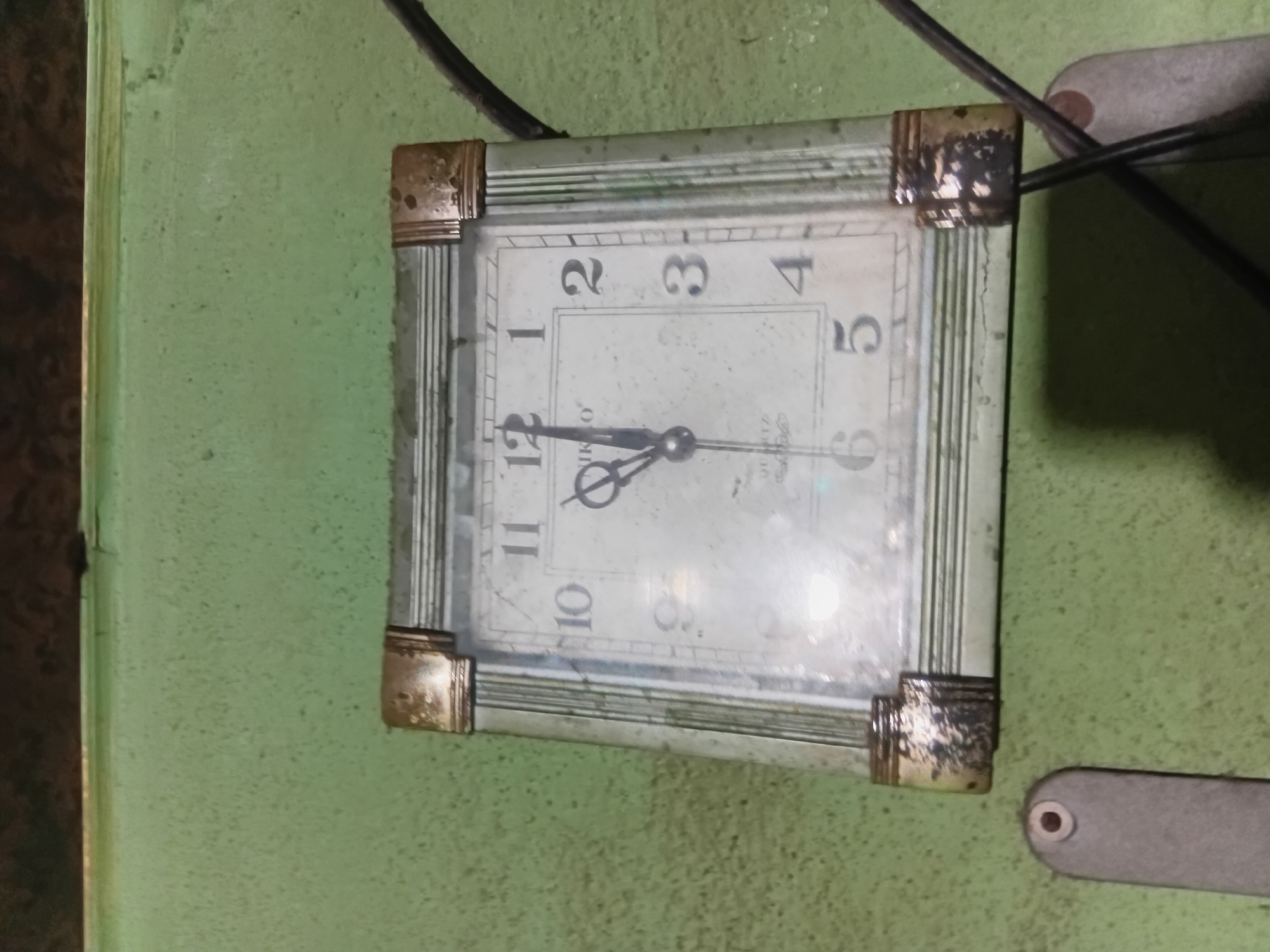The image shows a square clock with a white face and Roman numerals hanging on a textured, light green wall. The clock has a metallic frame with tarnished gold-colored corner accents. The hands of the clock are visible, with the hour hand pointing slightly before 12 and the minute hand pointing at 12. The word "QUARTZ" is visible below the center of the clock face. To the left and right of the clock are parts of a gray, oblong object with a circular indentation, possibly part of a mounting or fixture. Electrical wires are also visible to the right of the clock. The overall impression is of an interior setting, possibly in a home or public building in Nkpor, Nigeria, with the clock appearing to be old and somewhat weathered.