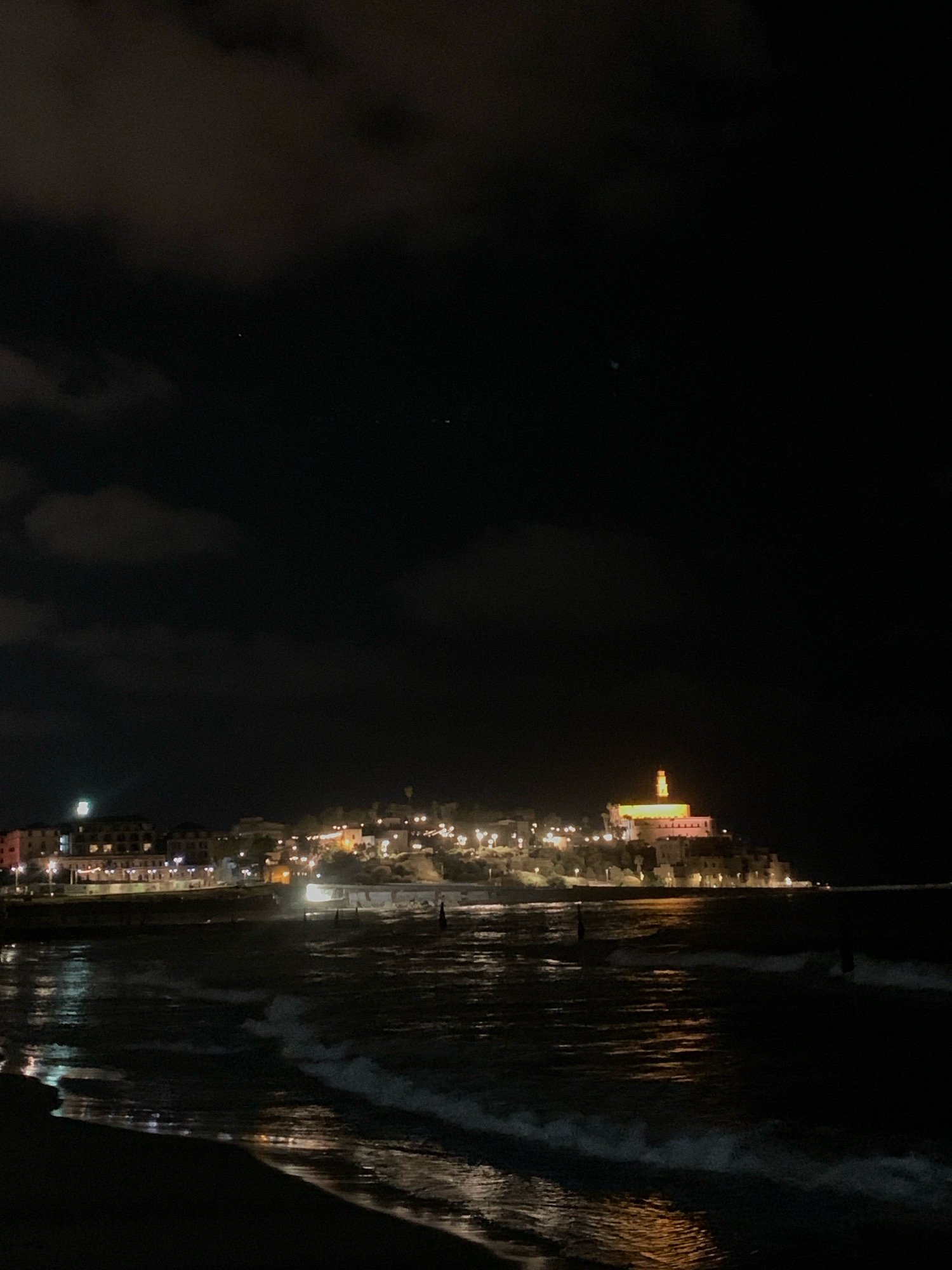 A nighttime, low-angle shot captures the Jaffa Port area of Tel Aviv, Israel, from a beach. The scene is dominated by the dark sky, with scattered clouds illuminated by ambient light. In the background, the historic city of Jaffa is visible on a hill, its buildings aglow with warm lights. A prominent, illuminated structure with a tower stands out on the hilltop. In the mid-ground, the port area features a breakwater with lights reflecting on the water. The foreground shows a sandy beach with gentle waves lapping at the shore. The water is dark, with reflections of the city lights shimmering on its surface. There are no discernible people or specific actions depicted.