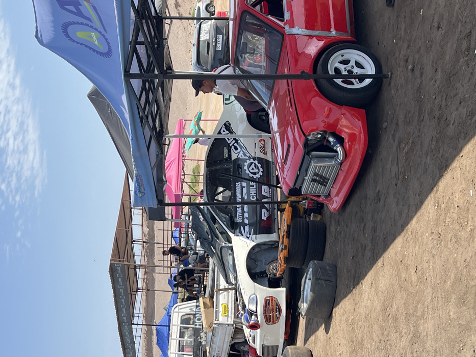 The image shows a collection of modified race cars parked in a pit area. A red car with visible front-mounted intercooler dominates the foreground. Behind it, a white car with sponsor decals and a black roll cage. People are seen under a shaded area. The scene has a dusty, outdoor atmosphere, likely a racetrack or similar event location.