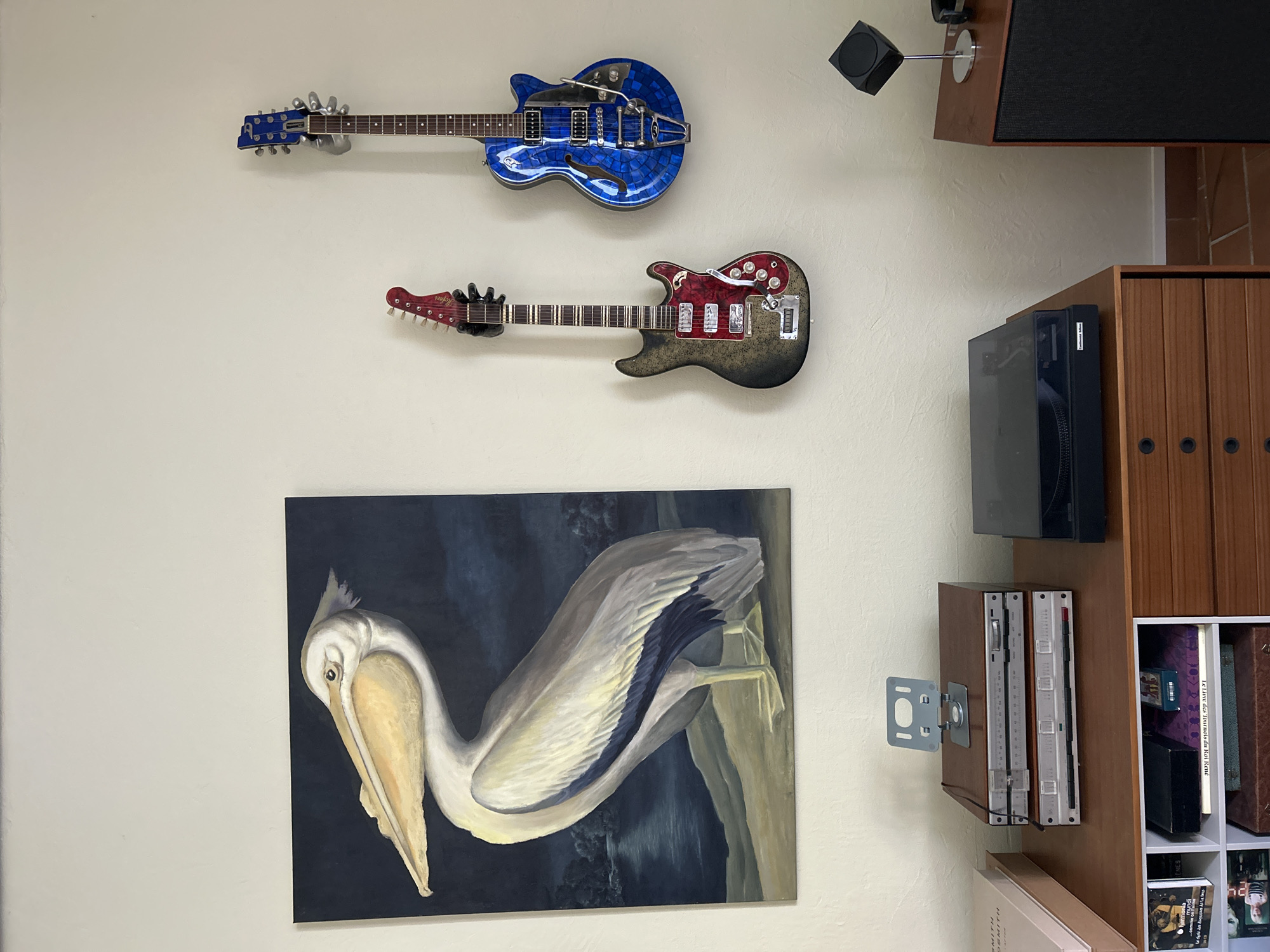 The image depicts an indoor scene with a wall adorned with two electric guitars and a painting of a pelican. The pelican painting, rendered in a realistic style, shows the bird standing on a sandy bank beside a body of water under a dark sky. To the right of the painting, two electric guitars are mounted on the wall. The guitar on the left is black and red with silver hardware, while the guitar on the right is a vibrant blue with a mosaic-like pattern and silver hardware.
Below the guitars, on a wooden dresser or cabinet, several electronic components are visible. This includes a stack of what appear to be audio receivers or amplifiers, with one unit featuring a stand holding a digital device. To the right of these components, a black turntable is placed on the surface of the dresser. The dresser itself is made of wood and has several drawers.
In the lower left corner of the image, part of a pink book is visible, and below that, a white cube shelf unit contains various items, including books and what appears to be a small black box. On the far right side of the image, a portion of a wooden speaker cabinet is visible, with a small, cube-shaped object on a stand placed on top of it. The lighting suggests an indoor environment with ambient light. The overall scene suggests a room designed for listening to music or displaying musical instruments. The location is stated as Saint-Marcel-lès-Sauzet, France, though there are no explicit visual cues within the image to confirm this specific location.