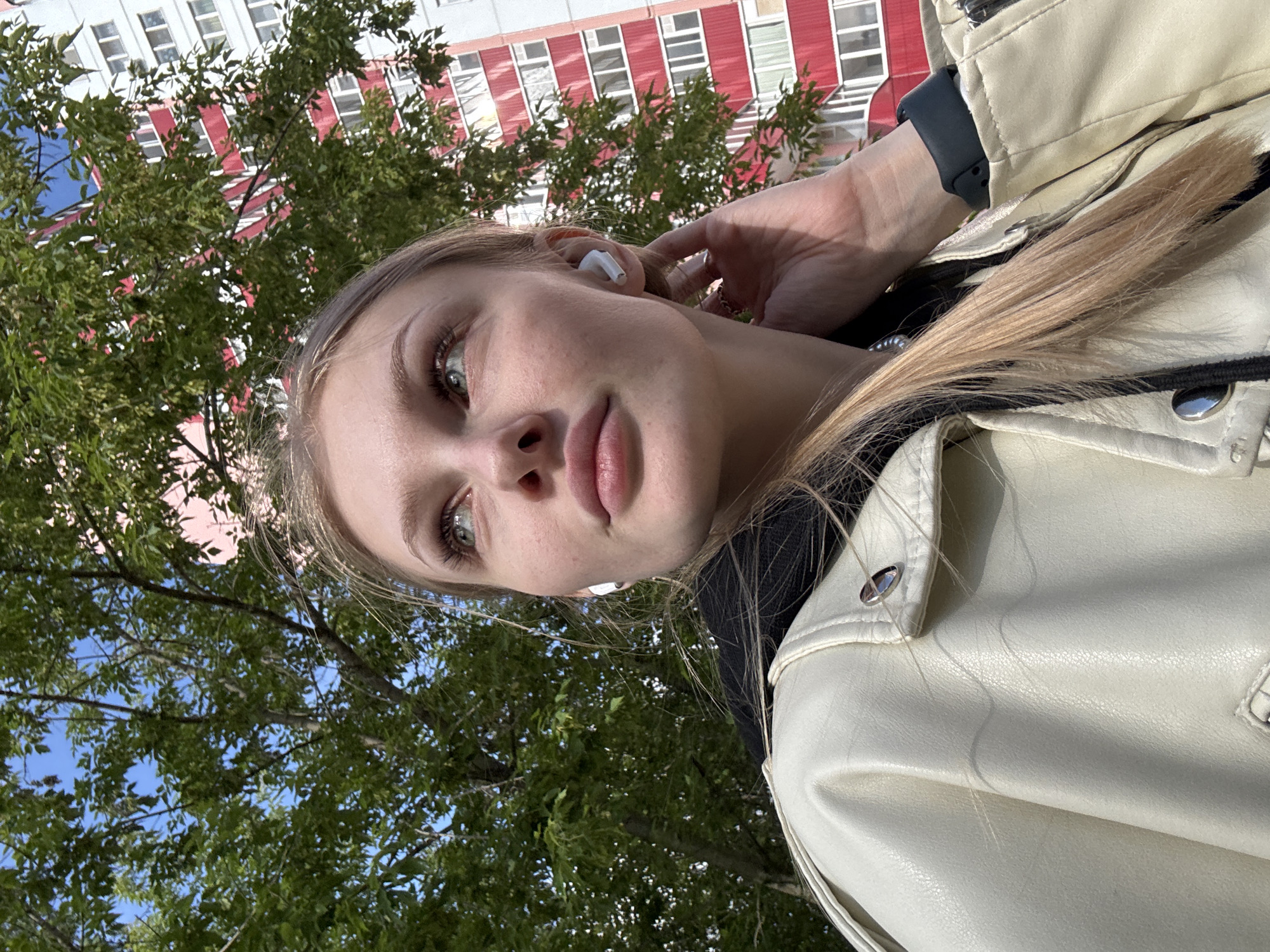 A young adult female is depicted in an outdoor, low-angle portrait, looking upwards and to her right. She has fair skin with visible freckles, prominent lips, and light-colored eyes, with long, blonde hair falling to her shoulder. She wears a cream-colored faux leather jacket over a dark hooded garment. White wireless earbuds are inserted into both ears, and a dark-banded smartwatch is visible on her left wrist. Her left hand is raised, with fingers touching the area near her left ear. The background features a dense canopy of green deciduous trees on the left, and a multi-story residential building on the right. The building is white with distinct red horizontal banding between floors and multiple windows and balconies, consistent with urban architecture in Perm, Russia. The scene is illuminated by natural daylight under a clear blue sky.