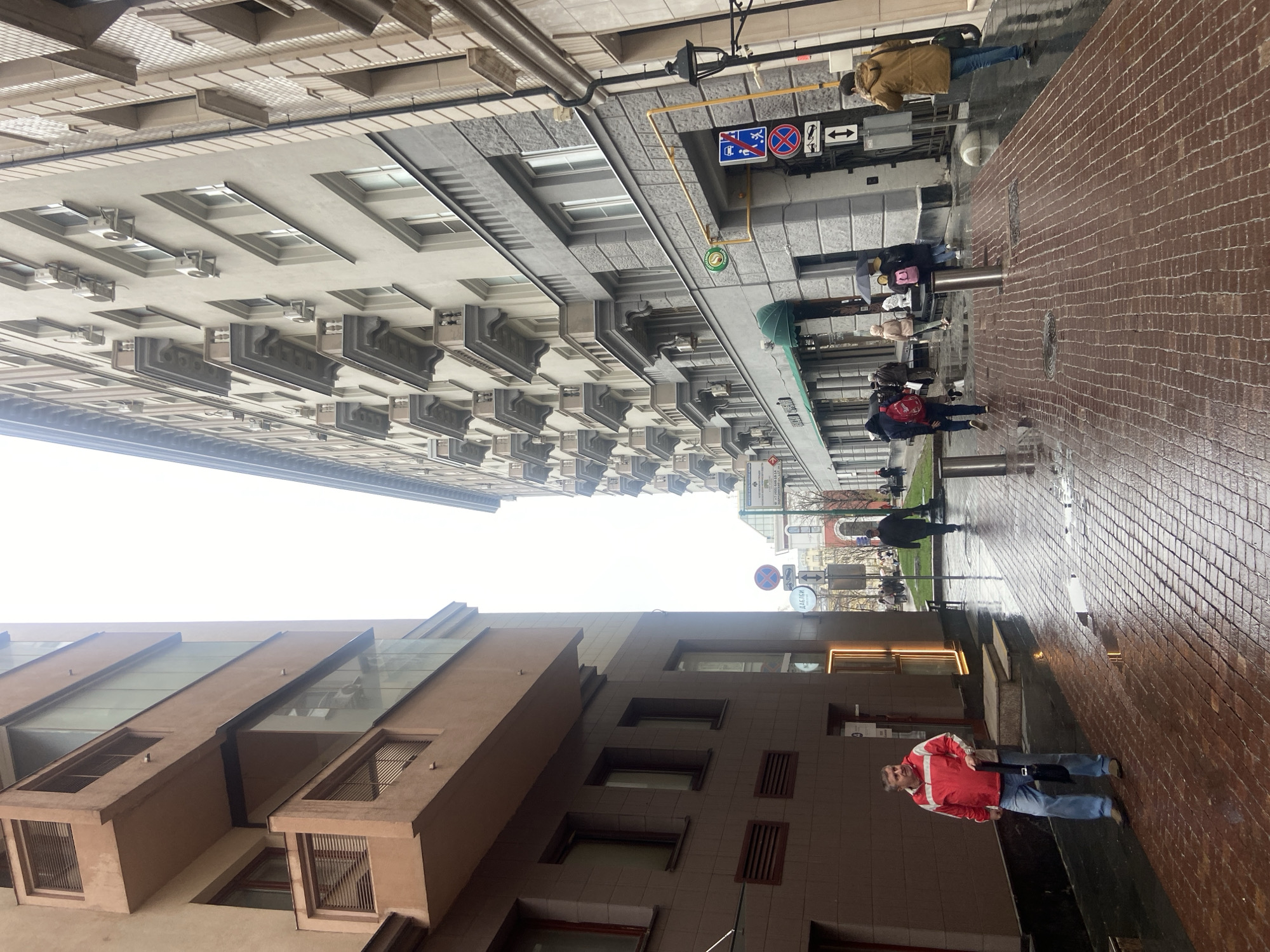 Here is a description of the image:
The image shows a street scene in Moscow, Russia, taken on a wet day. The perspective is looking up between buildings on either side of the street. The buildings are tall and have different architectural styles. The building on the left is modern with geometric windows and some glass sections. The building on the right is older and more ornate, with numerous air conditioning units mounted below the windows.
The street is paved with red bricks and is wet, reflecting the buildings and sky. Several pedestrians are walking along the sidewalk, holding umbrellas. There is a man in a red jacket standing on the right sidewalk. Street signs are visible, including a no-parking sign and a sign indicating direction for cars. The sky is overcast, and the lighting is subdued. The overall impression is of a typical rainy day in a bustling urban environment.
