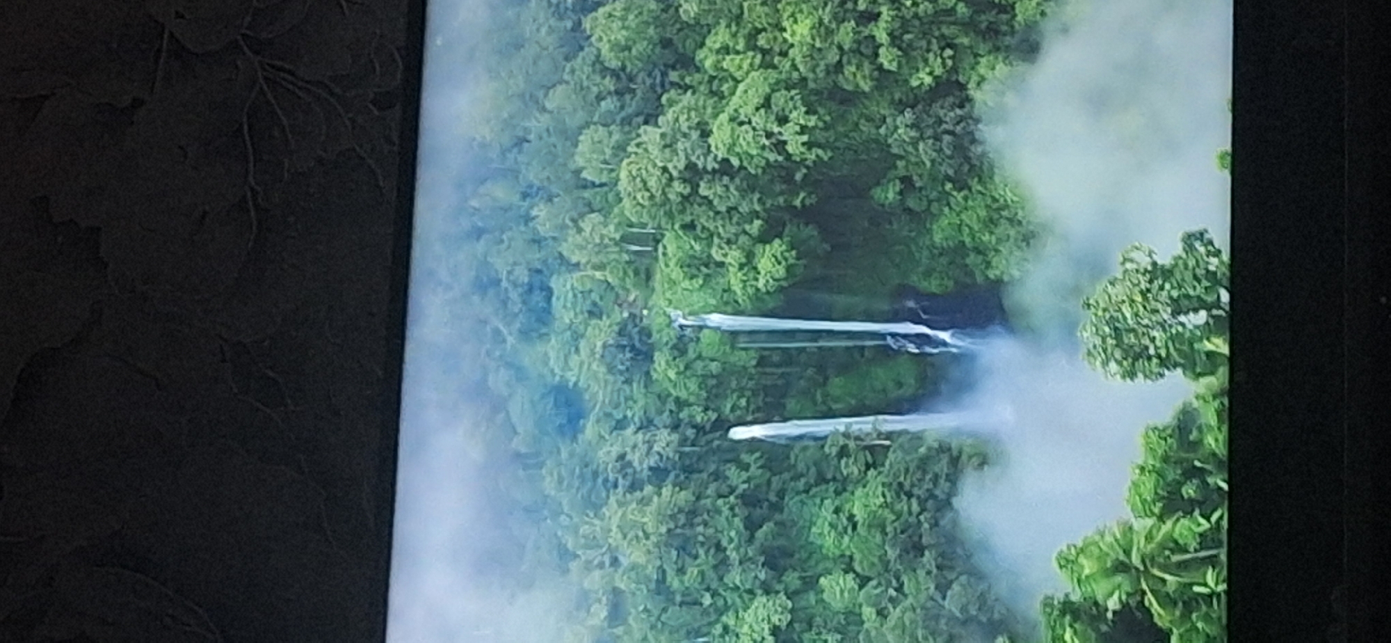 The image displays an aerial view of a lush, dense jungle with two prominent waterfalls cascading down a cliff face. The waterfalls are partially obscured by mist, creating a dramatic and ethereal atmosphere. The surrounding jungle is teeming with vibrant green foliage, suggesting a tropical or subtropical environment. The scene is characterized by its natural beauty and the raw power of the cascading water. There are no people or man-made structures visible in the image. The location in the city of Bila Tserkva, Ukraine, is not ascertainable from the provided image.